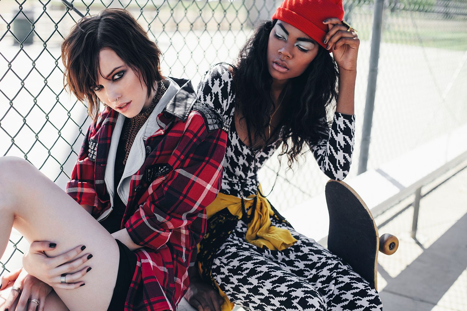 Band members in red plaid shirts sit against chain link fence holding guitars and instruments in outdoor setting.