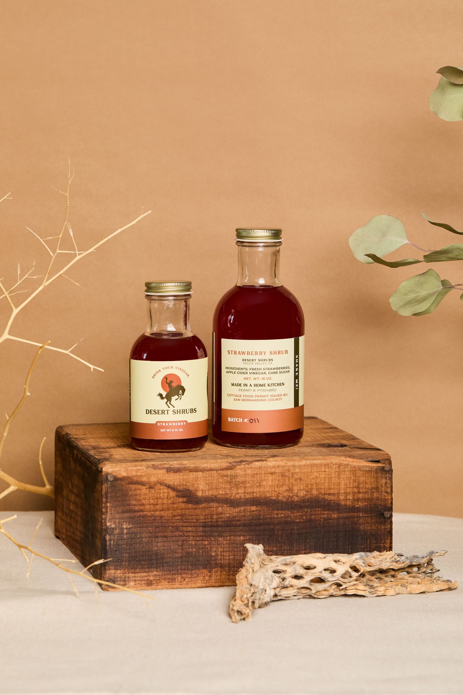 Two glass bottles of Desert Shrub syrup displayed on a rustic wooden box with eucalyptus and branches.