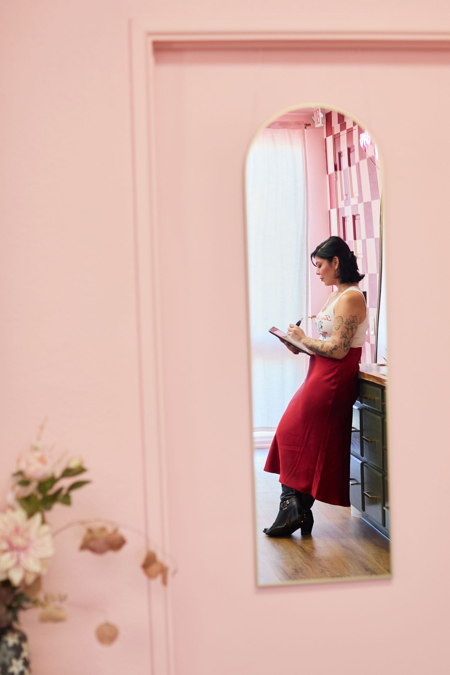 Woman in red skirt writing notes in a pink-walled boutique shop, viewed through an arched doorway.