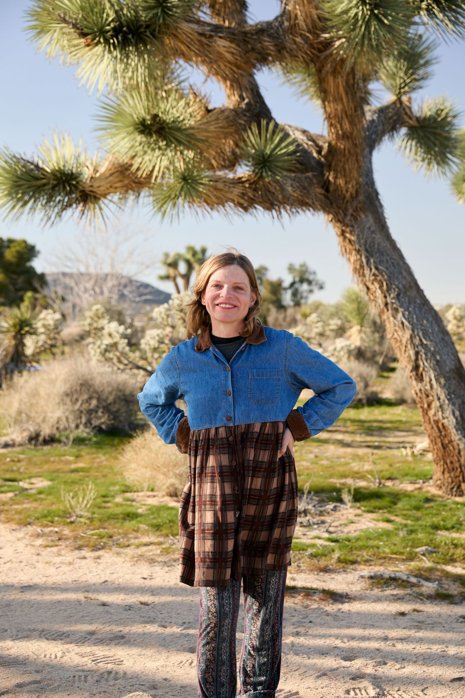 A person wearing a denim jacket and plaid shirt stands in a desert landscape with Joshua trees and mountains in the background.