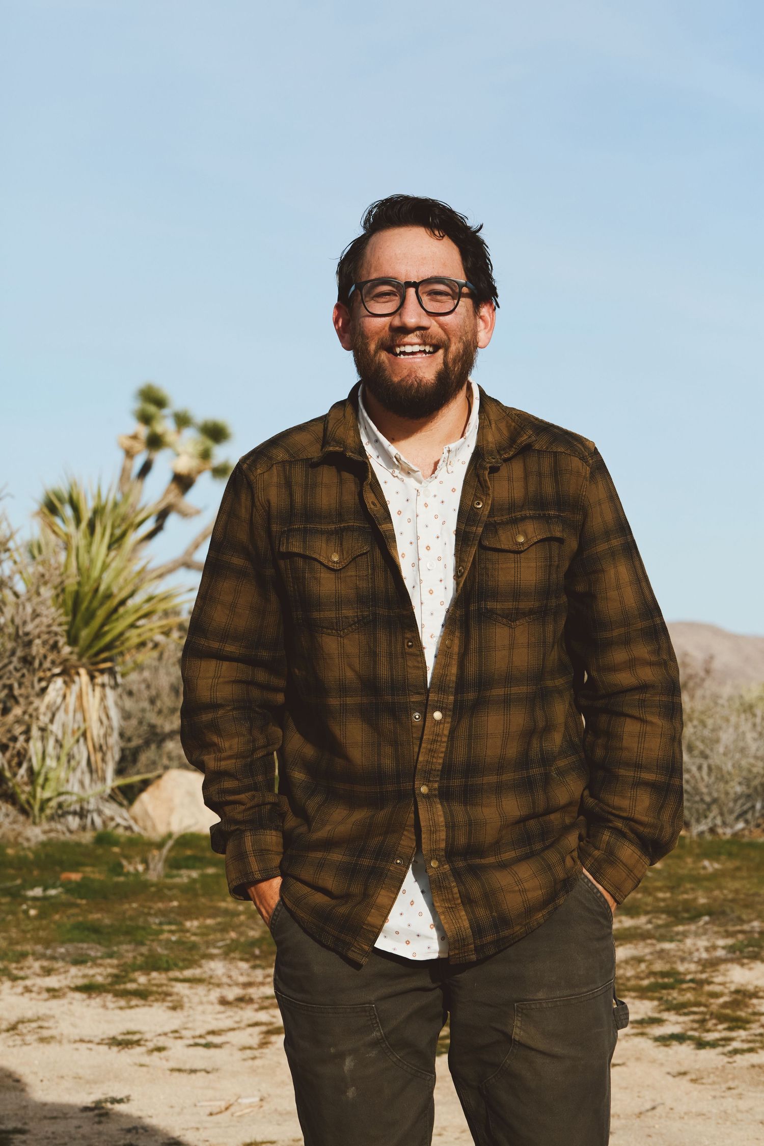 A person wearing glasses and a brown plaid flannel jacket smiles while standing outdoors with desert plants in the background.
