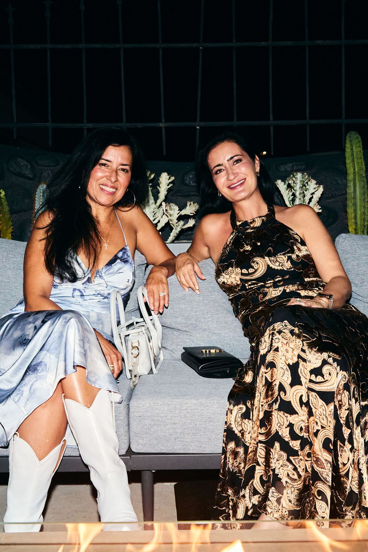 Two women in elegant dresses sitting on a grey couch with decorative cactus plants in the background.