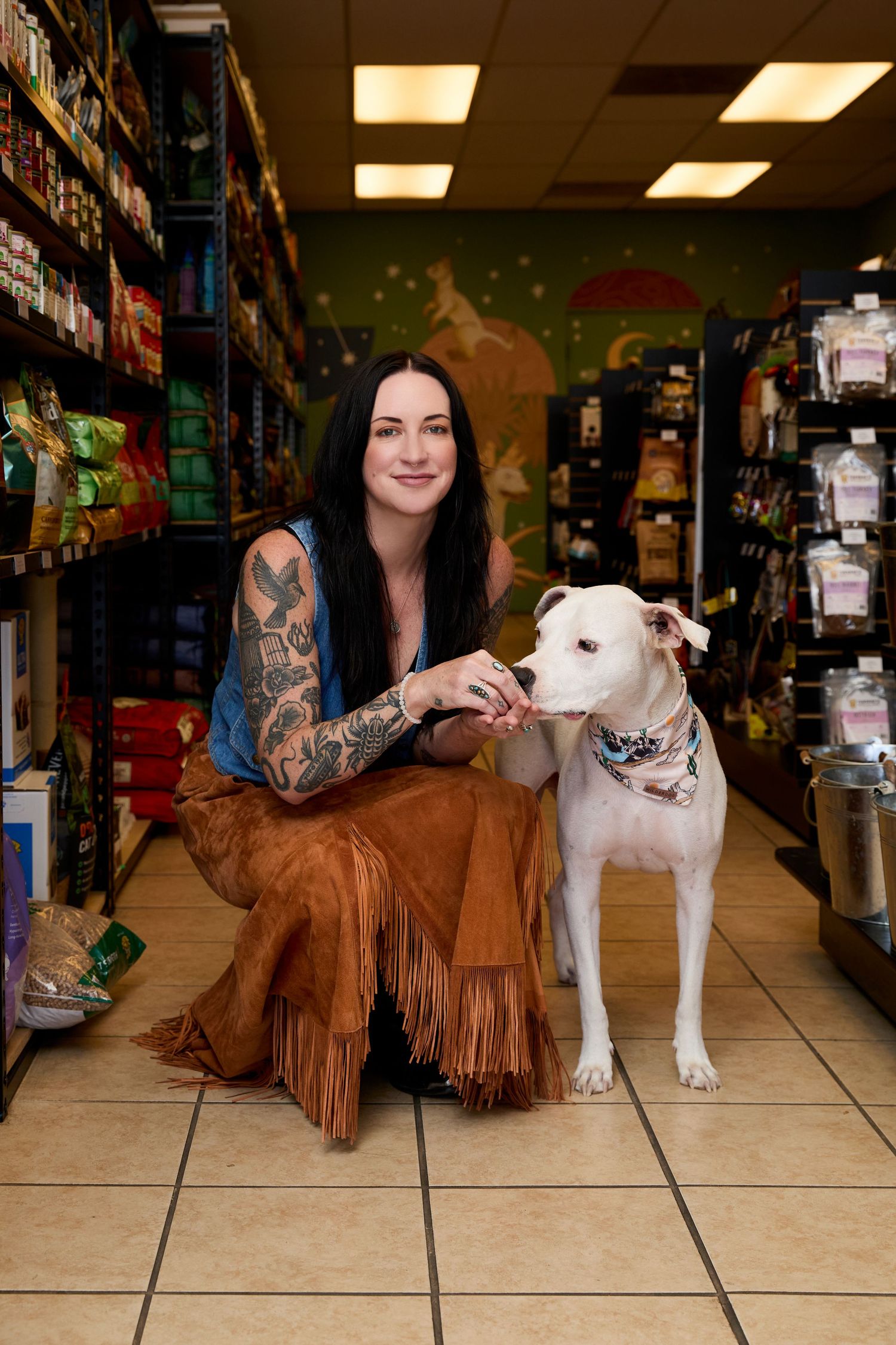 A white pit bull puppy poses with someone in a brown fringe jacket on tiled floor at a pet store.