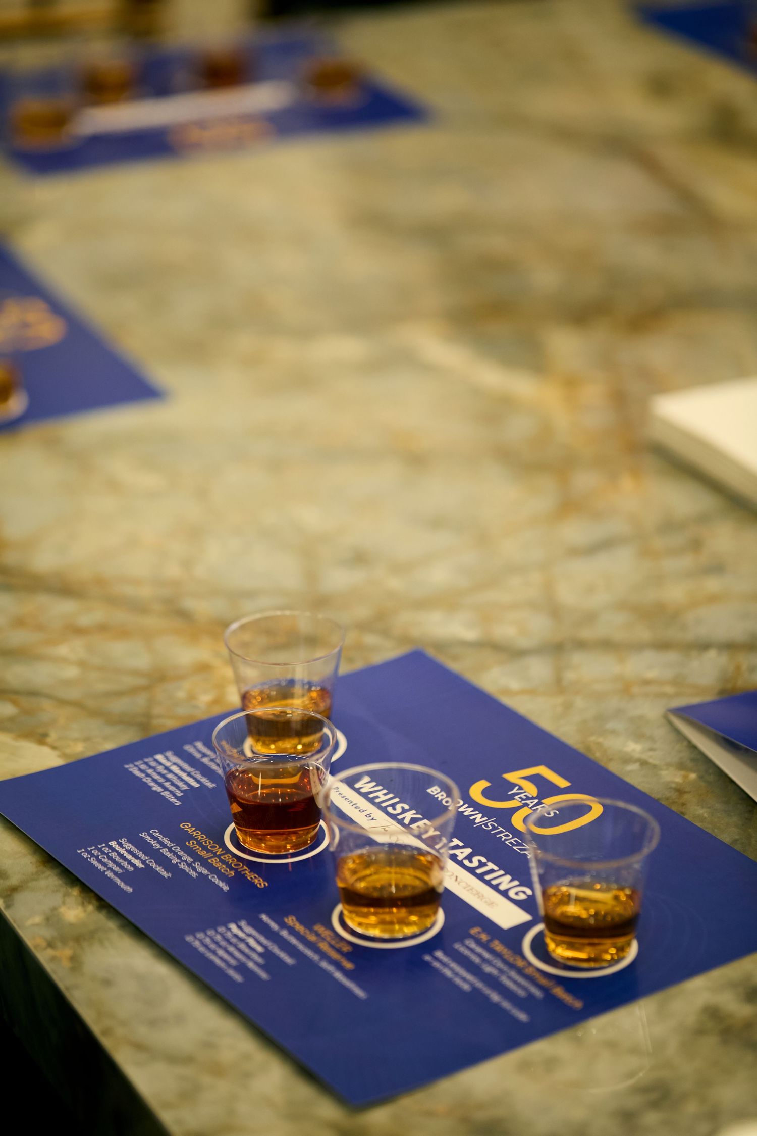 Small glasses of amber-colored whiskey arranged on blue menu cards against a marble surface.