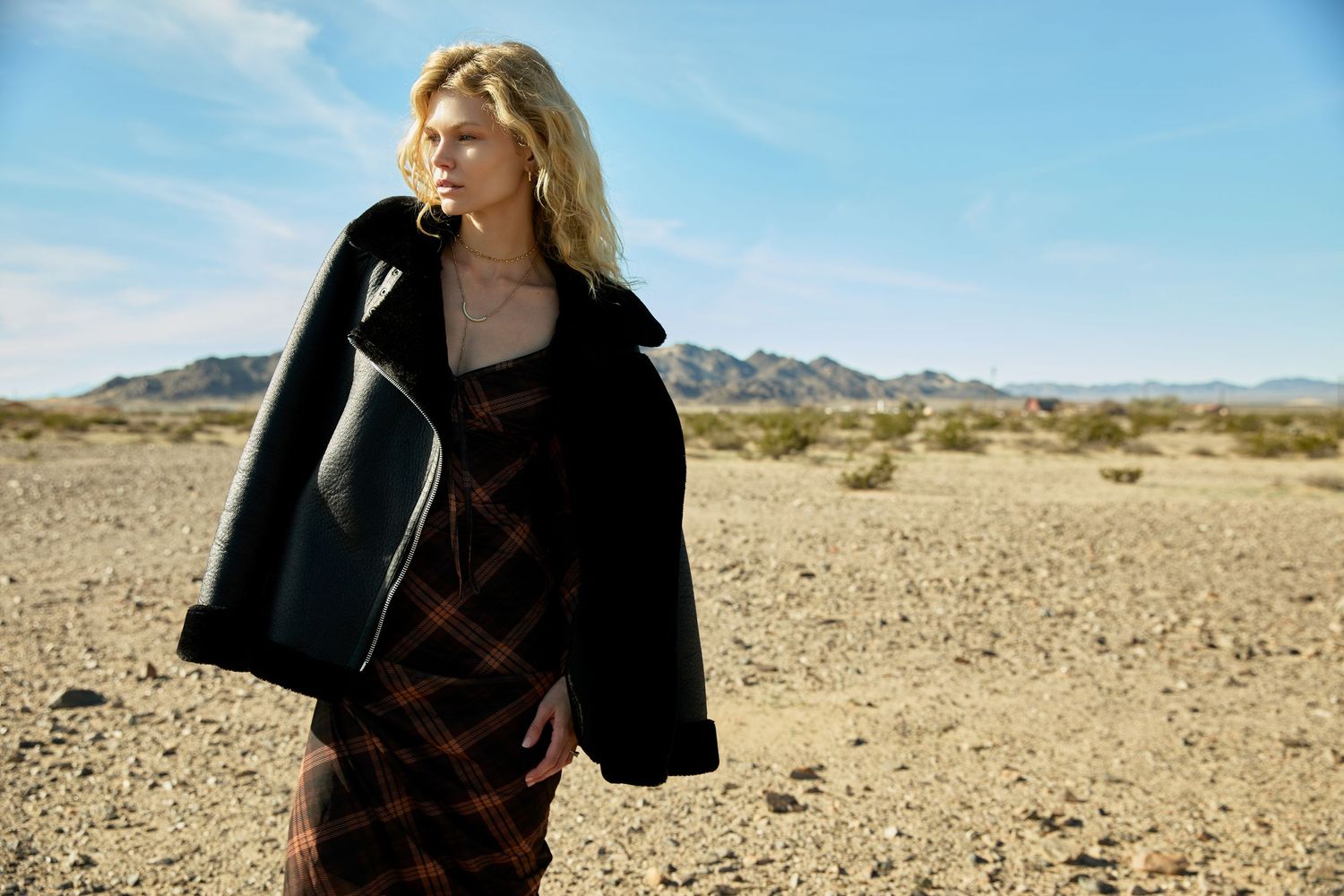 Figure in black jacket poses against desert landscape with mountains in background under bright blue sky.