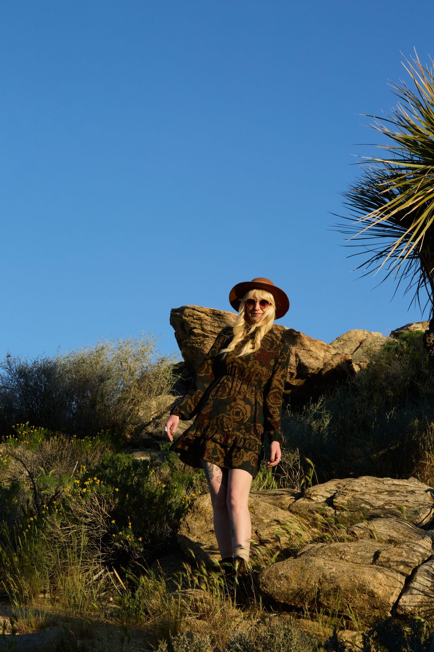 Blonde woman in brown patterned dress and hat poses on desert rocks with blue sky and cacti.