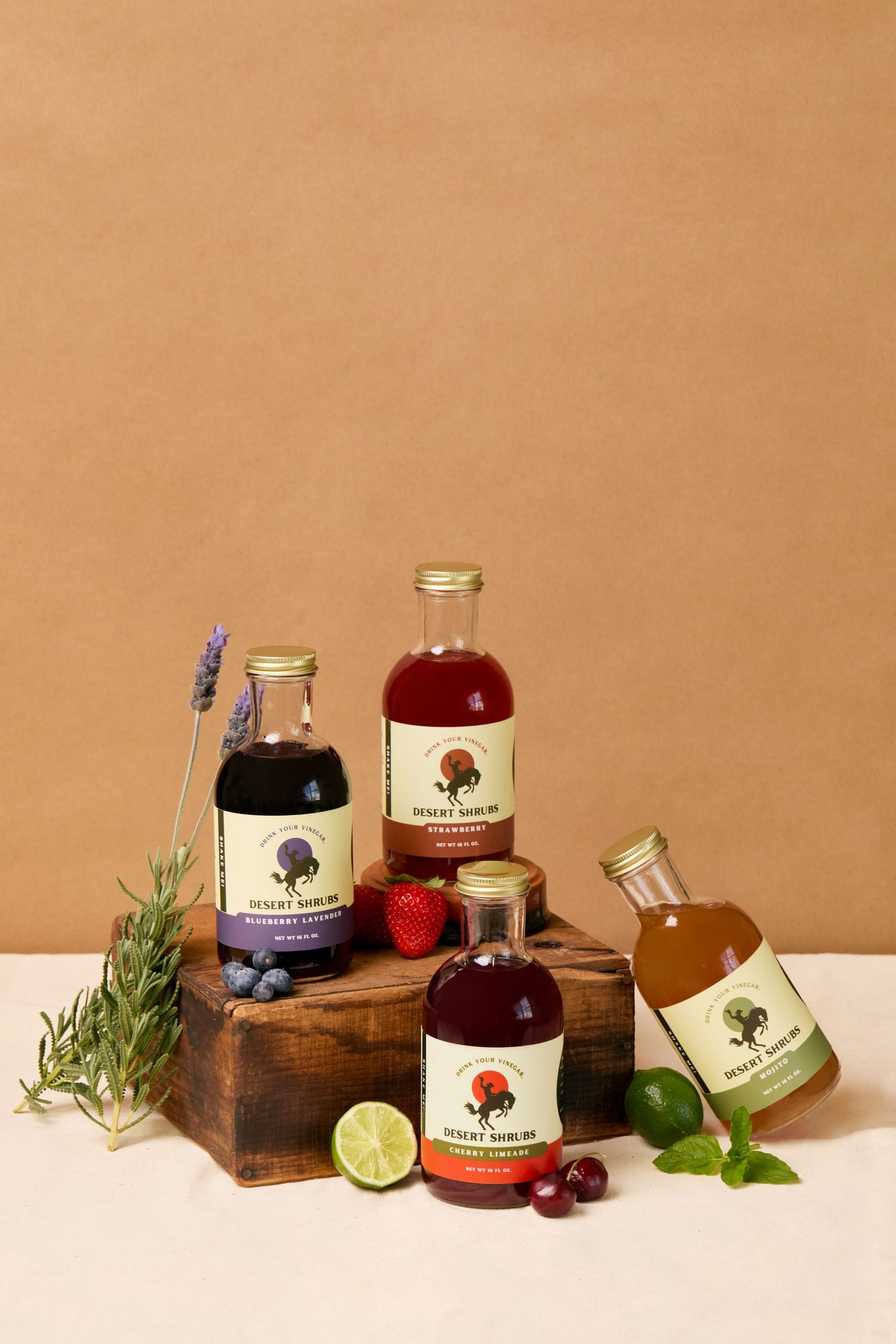 Four Desert Shrub craft cocktail syrup bottles displayed on a wooden block with fresh fruits and herbs on a tan background.