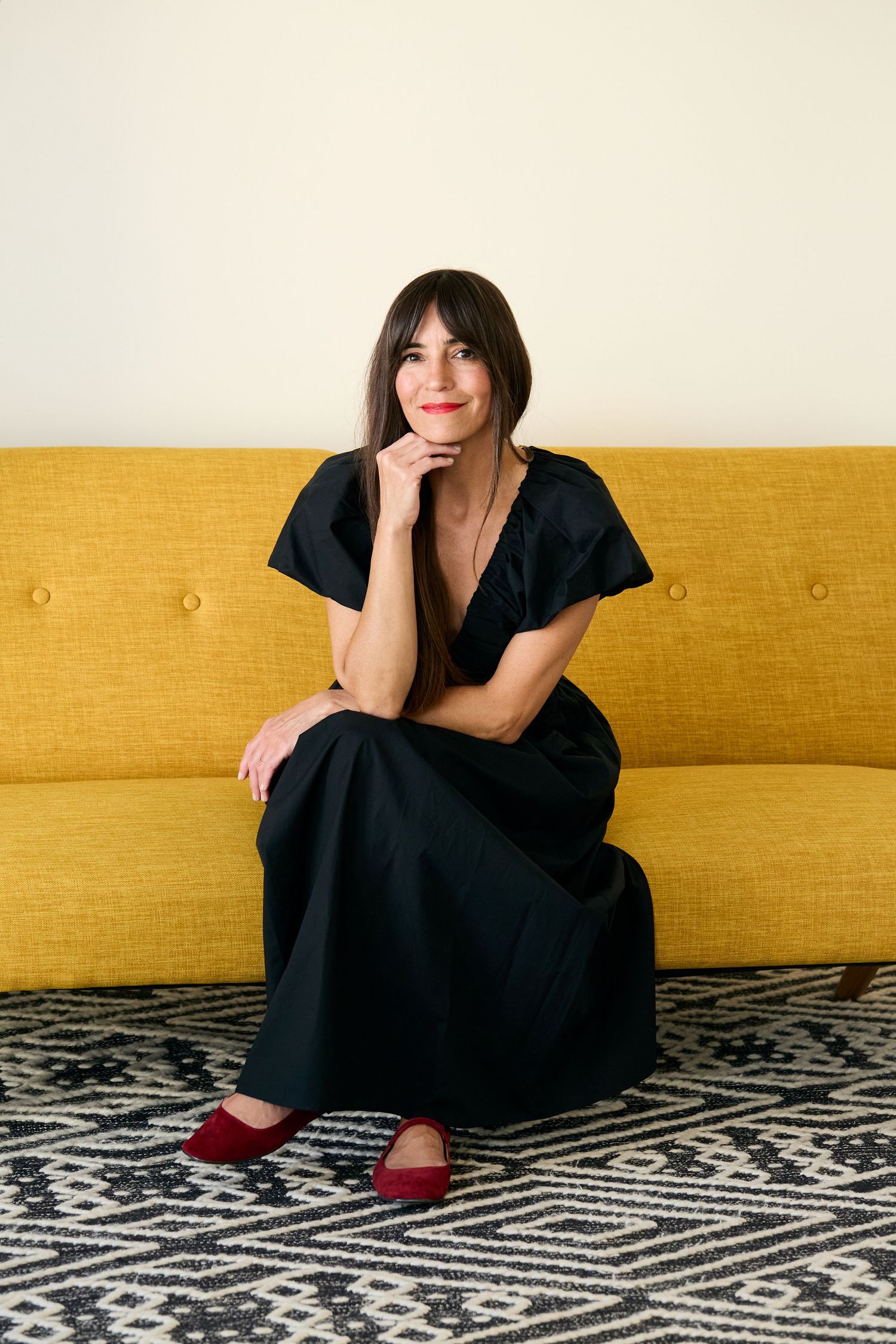 Person in black dress reclining on yellow couch with geometric patterned rug in modern minimalist setting.