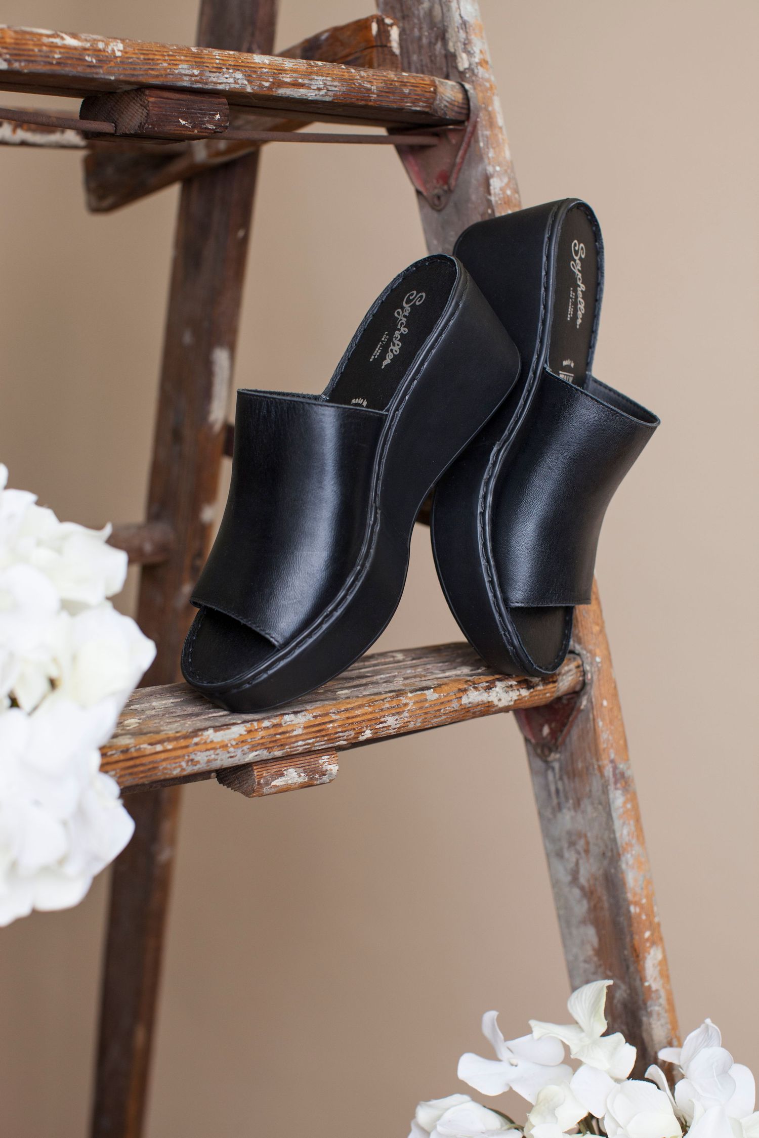 Black leather mule sandals displayed on rustic wooden ladder steps with white floral decorations.