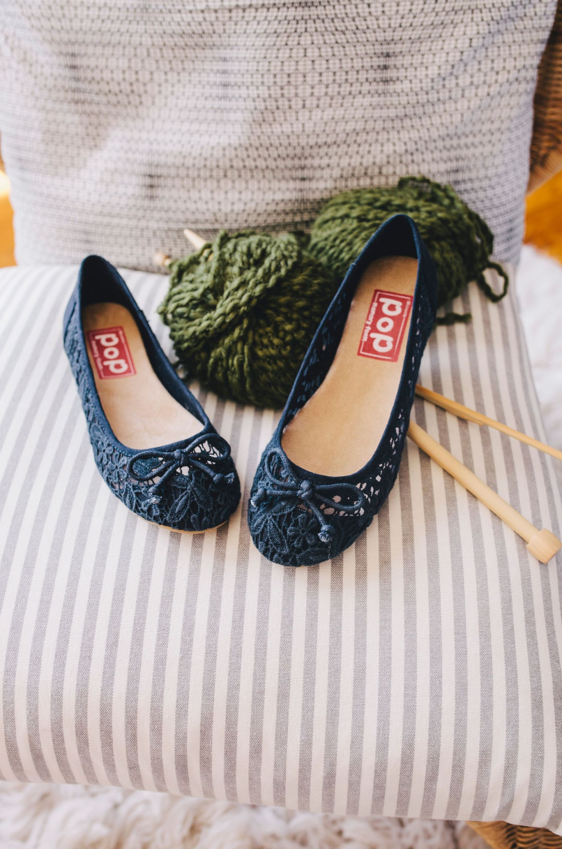 Navy blue lace flats with POP branding displayed on striped cushion with green knitting.