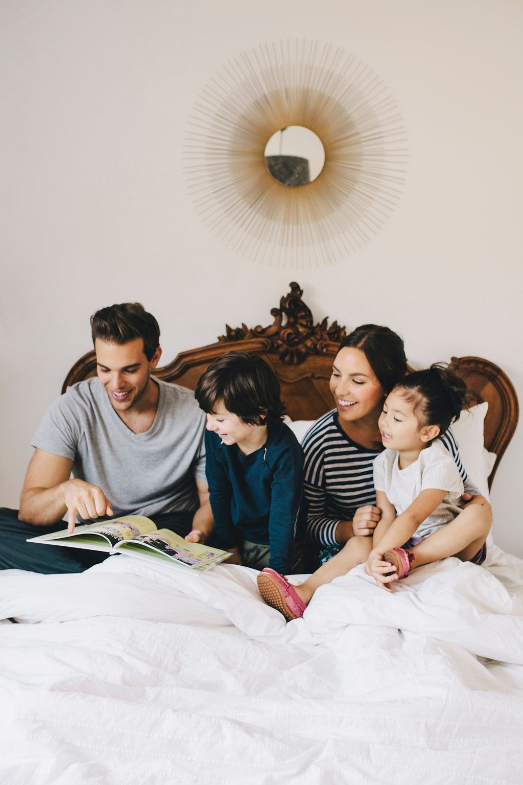 Family reading together on bed beneath decorative sunburst mirror.