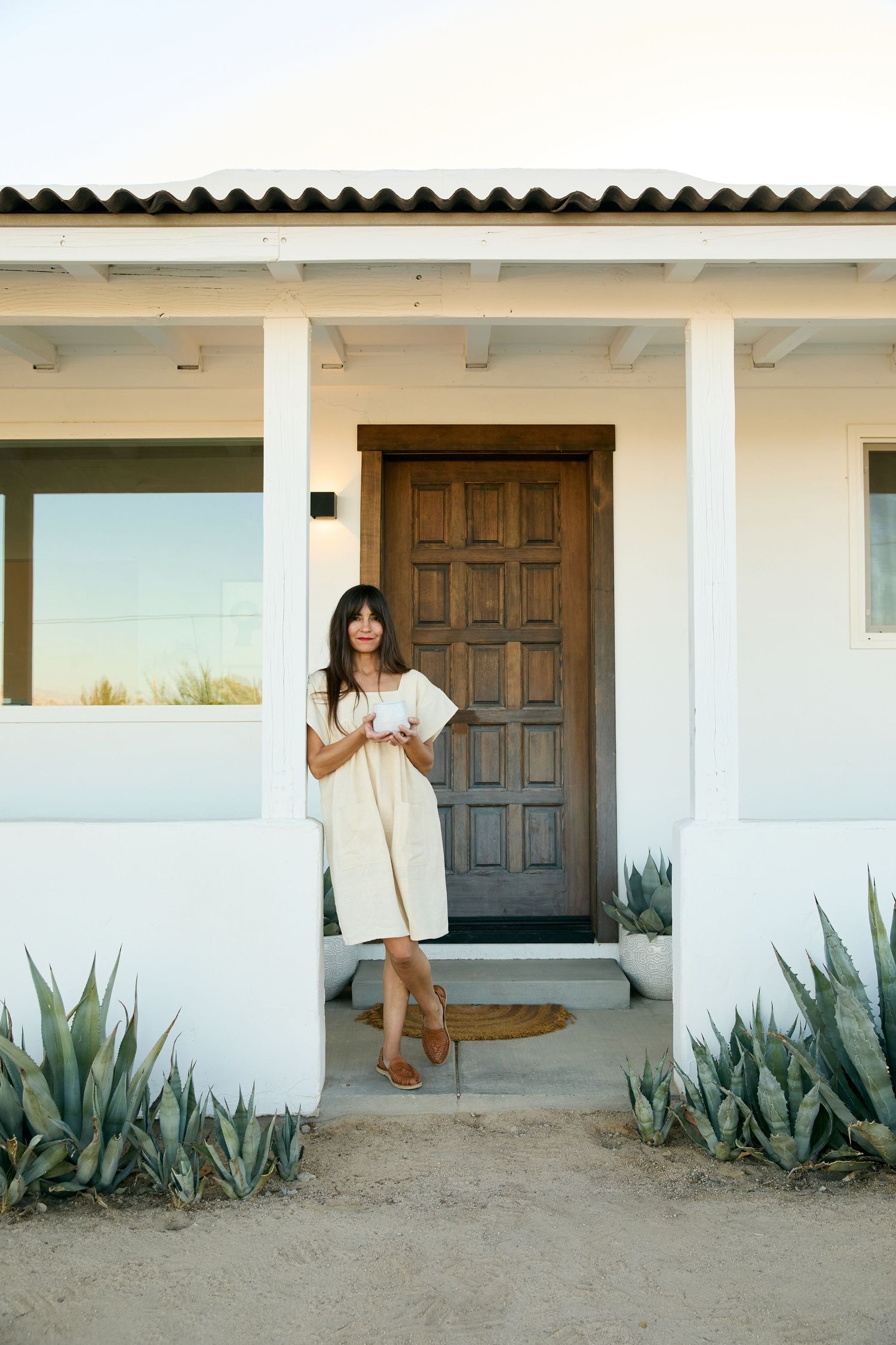 A figure stands in front of a white Spanish-style house with wooden door and agave plants in a desert landscape.