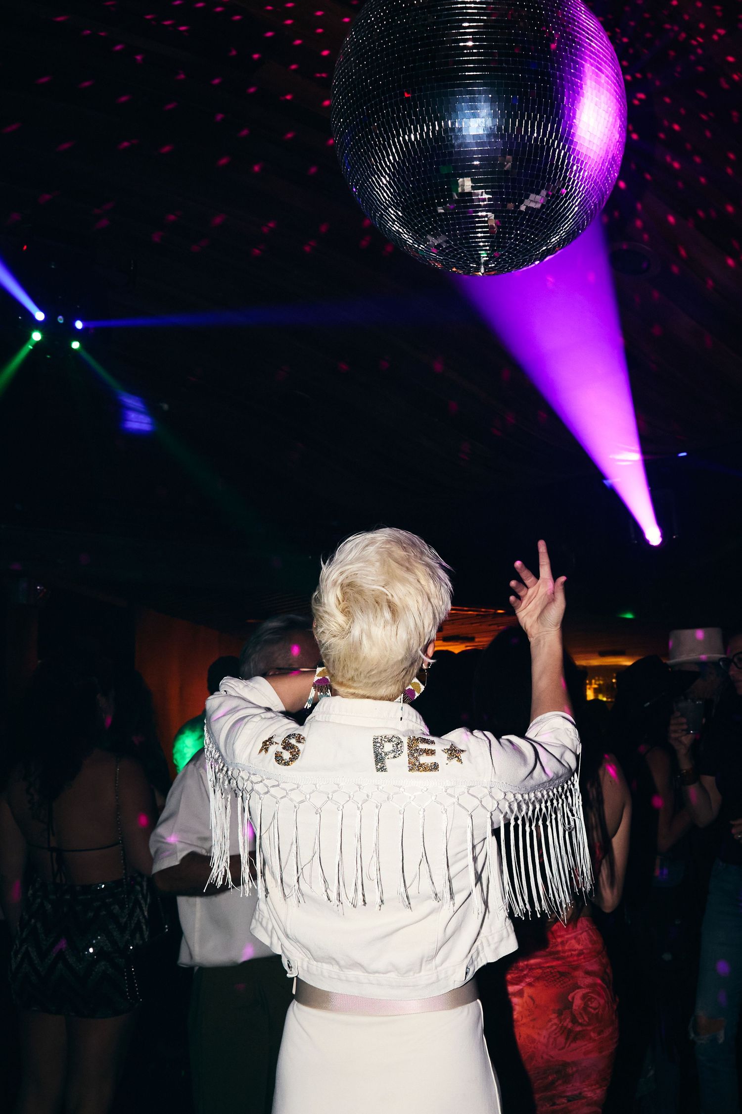 Person in white fringe jacket dancing under a disco ball with purple lighting at a nightclub party.