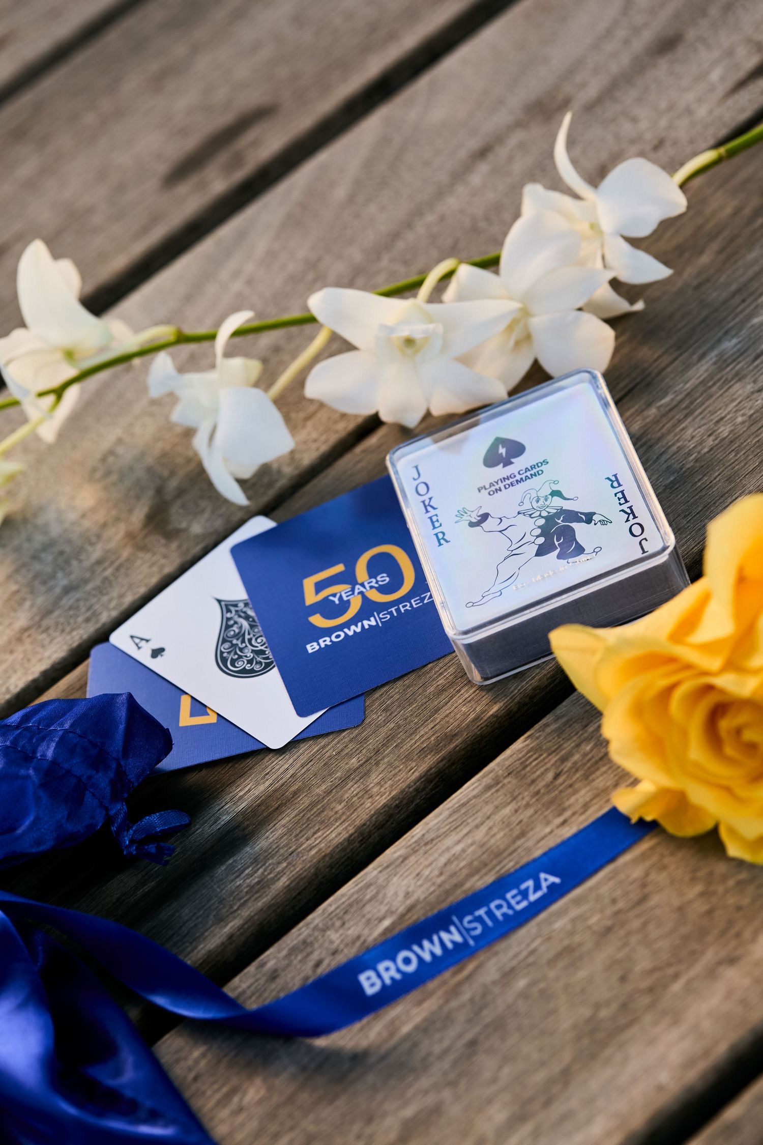 Playing cards and flowers arranged on rustic wooden deck with blue ribbon and white blossoms.
