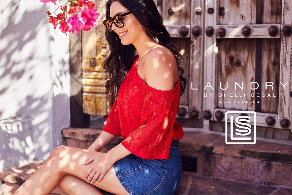 Woman in red off-shoulder top and denim shorts sitting beside flowering bushes.