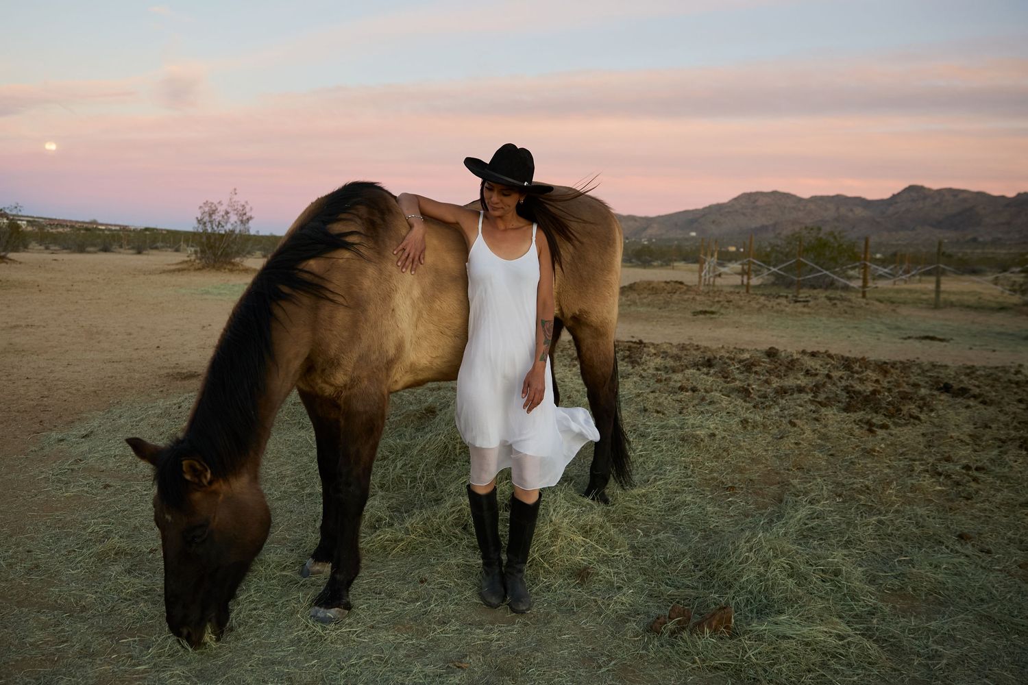 Woman in white dress and cowboy hat stands beside a grazing horse at sunset in a desert landscape.
