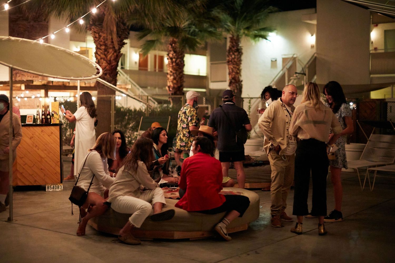 People gather at an outdoor evening market with palm trees and string lights creating an ambient atmosphere.