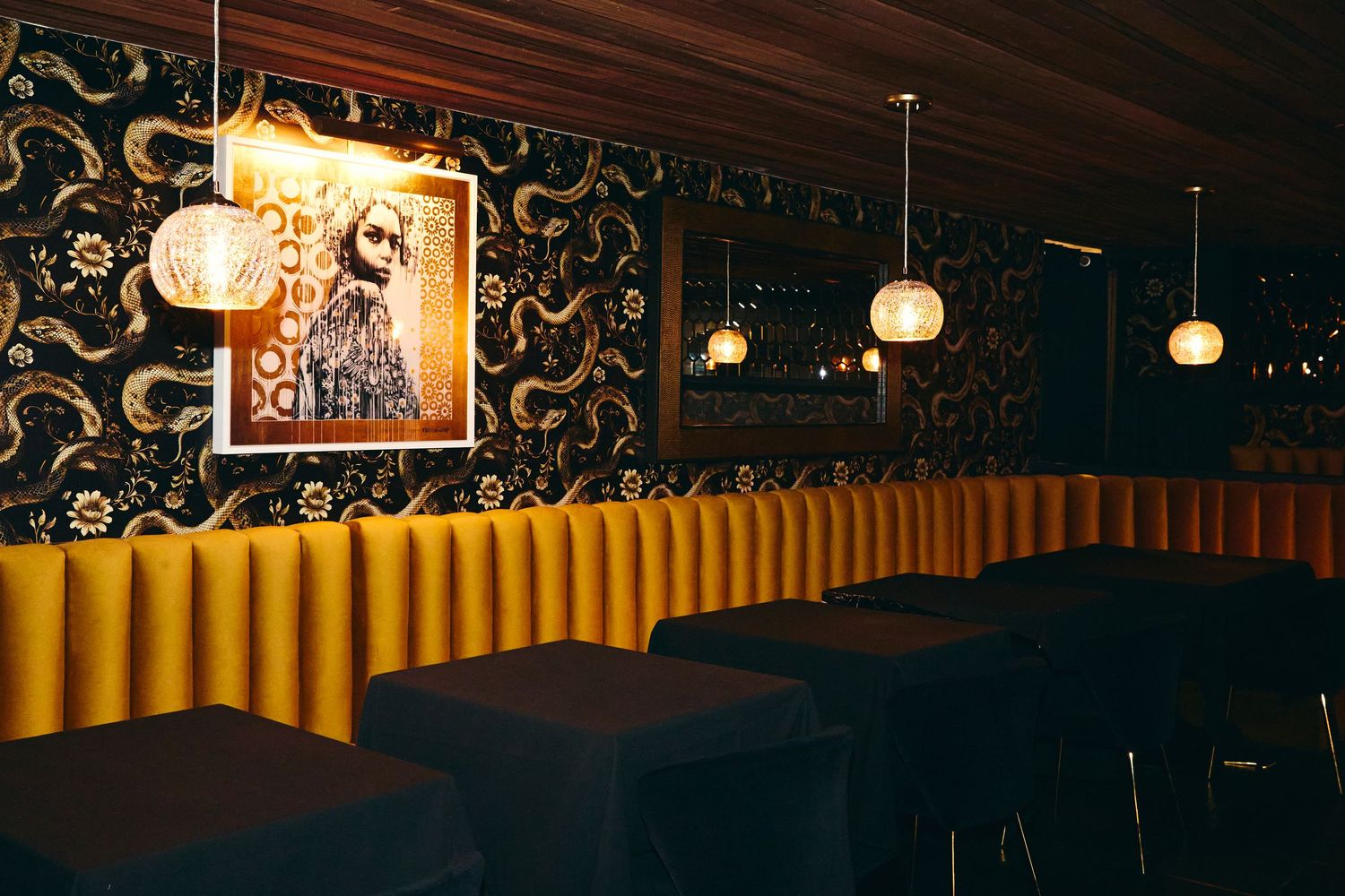 Retro lounge interior with yellow booth seating, dark wallpaper, and hanging pendant lights creating moody ambiance.