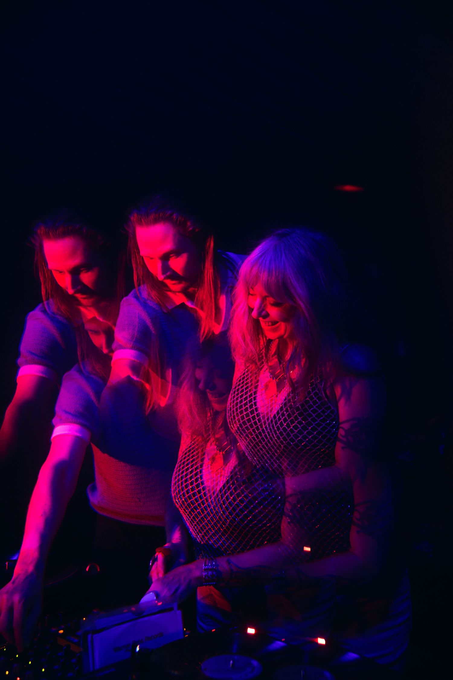 People dancing in a dark club environment lit with vibrant red and blue lights near DJ equipment.