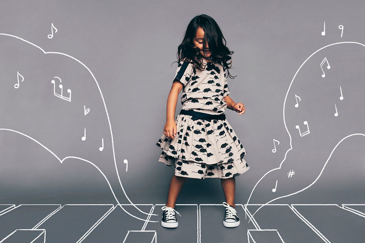 A child in a polka dot dress and sneakers dances against a gray wall decorated with chalk-drawn musical notes.