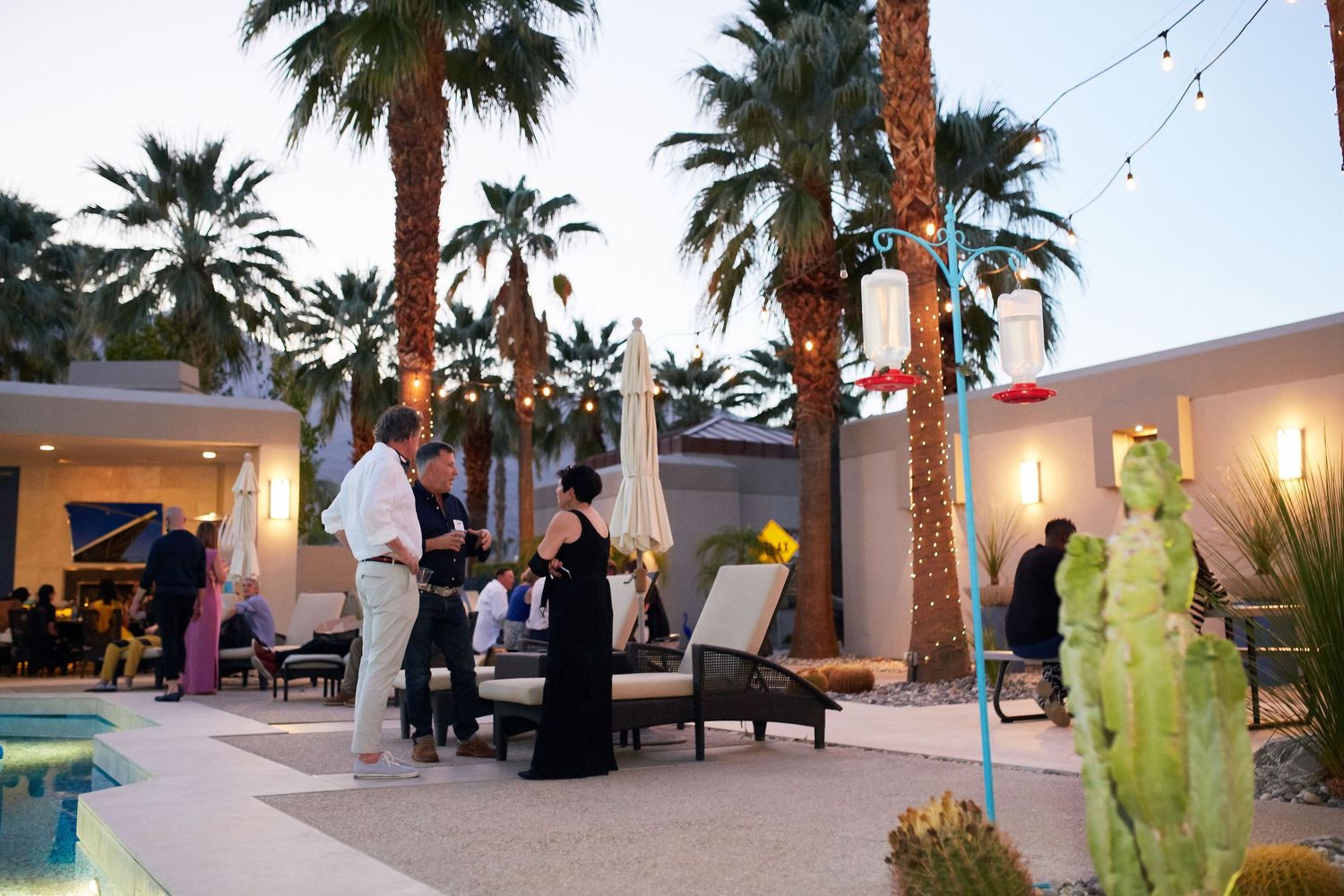 People gathered at a modern outdoor patio with palm trees and string lights creating an elegant evening ambiance by the pool.