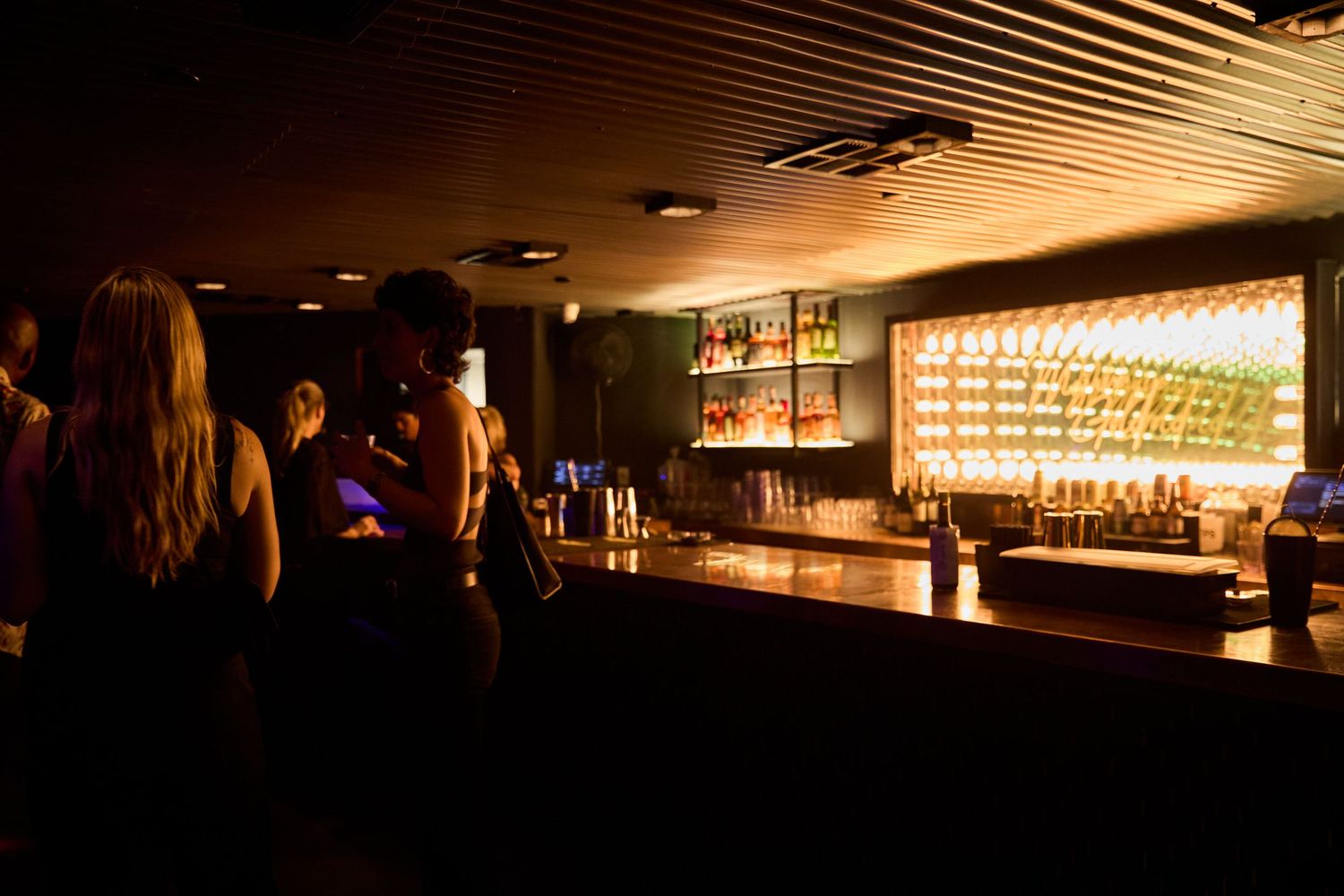 A dimly lit upscale bar interior with backlit shelves of bottles and customers seated at the counter.