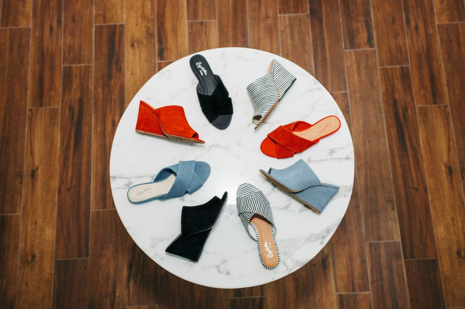 Colorful collection of summer sandals arranged in a circle on hardwood floor.
