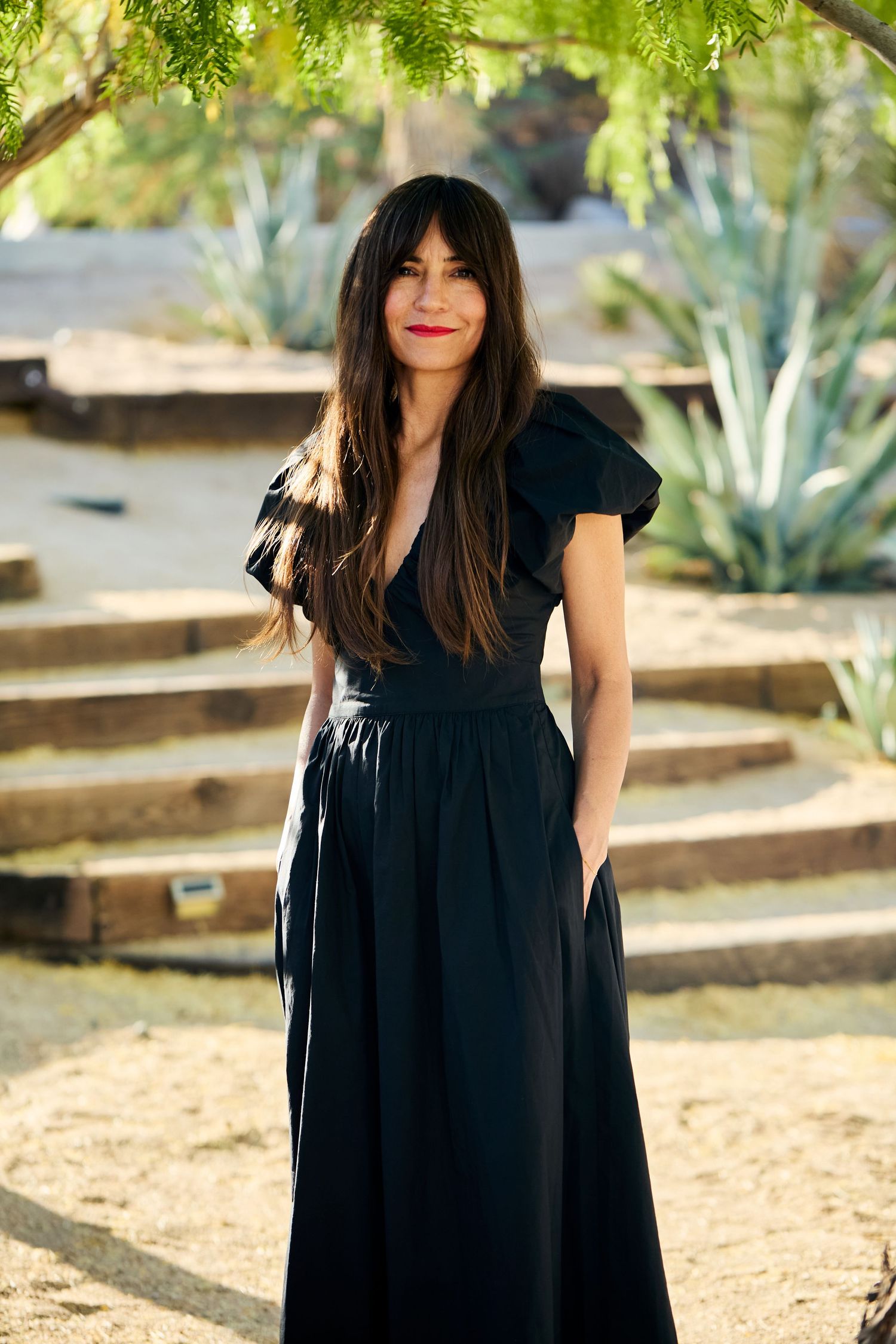 A person in a flowing black dress stands in a sunny garden setting with agave plants and trees in the background.