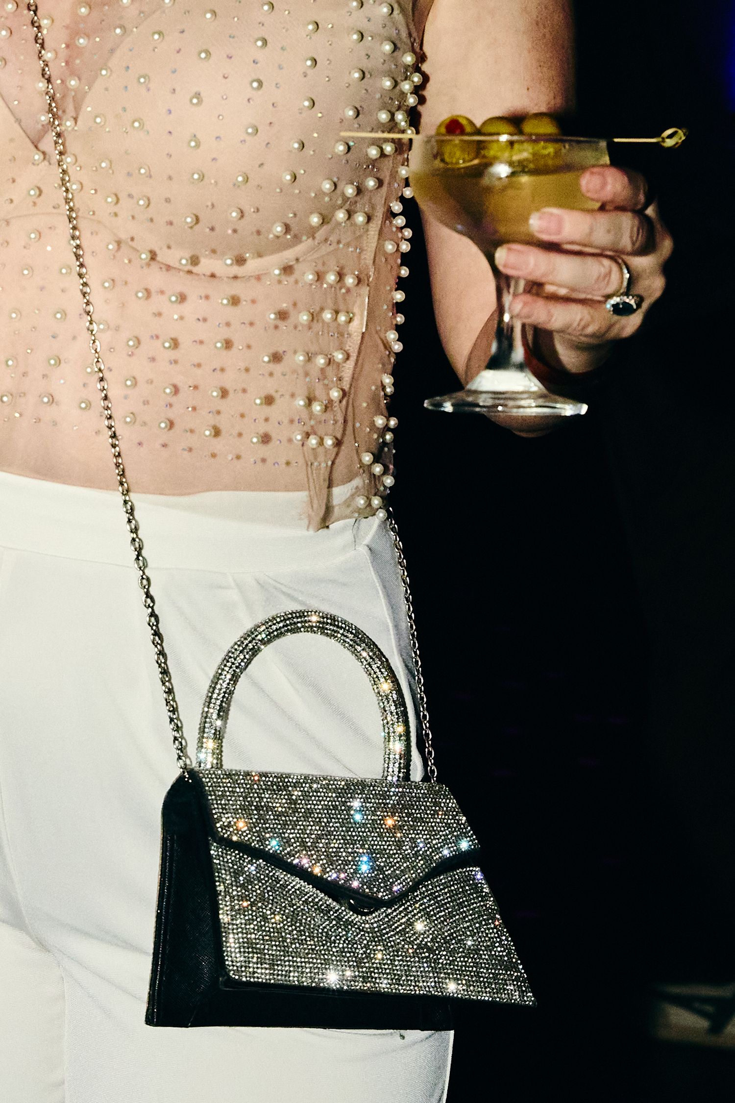 A sparkly silver handbag on a chain strap is shown next to a hand holding a martini glass with an olive.