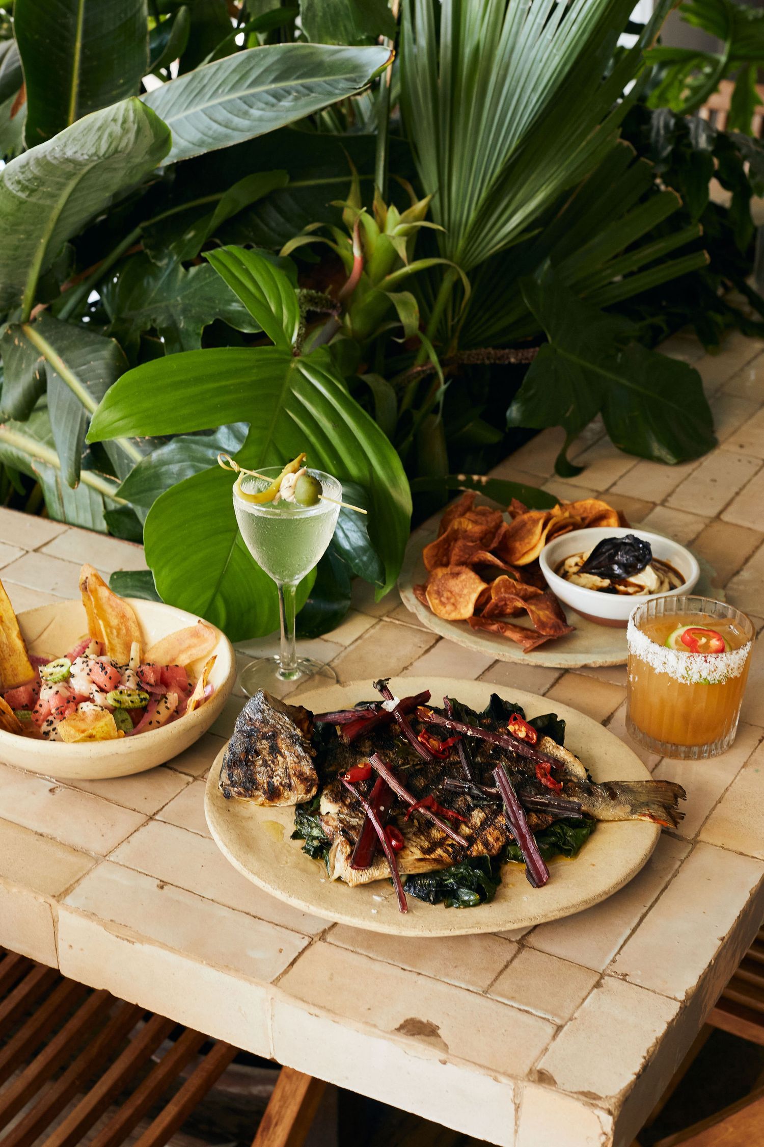 Tropical dining spread with grilled meats, cocktails and side dishes among lush green plants.