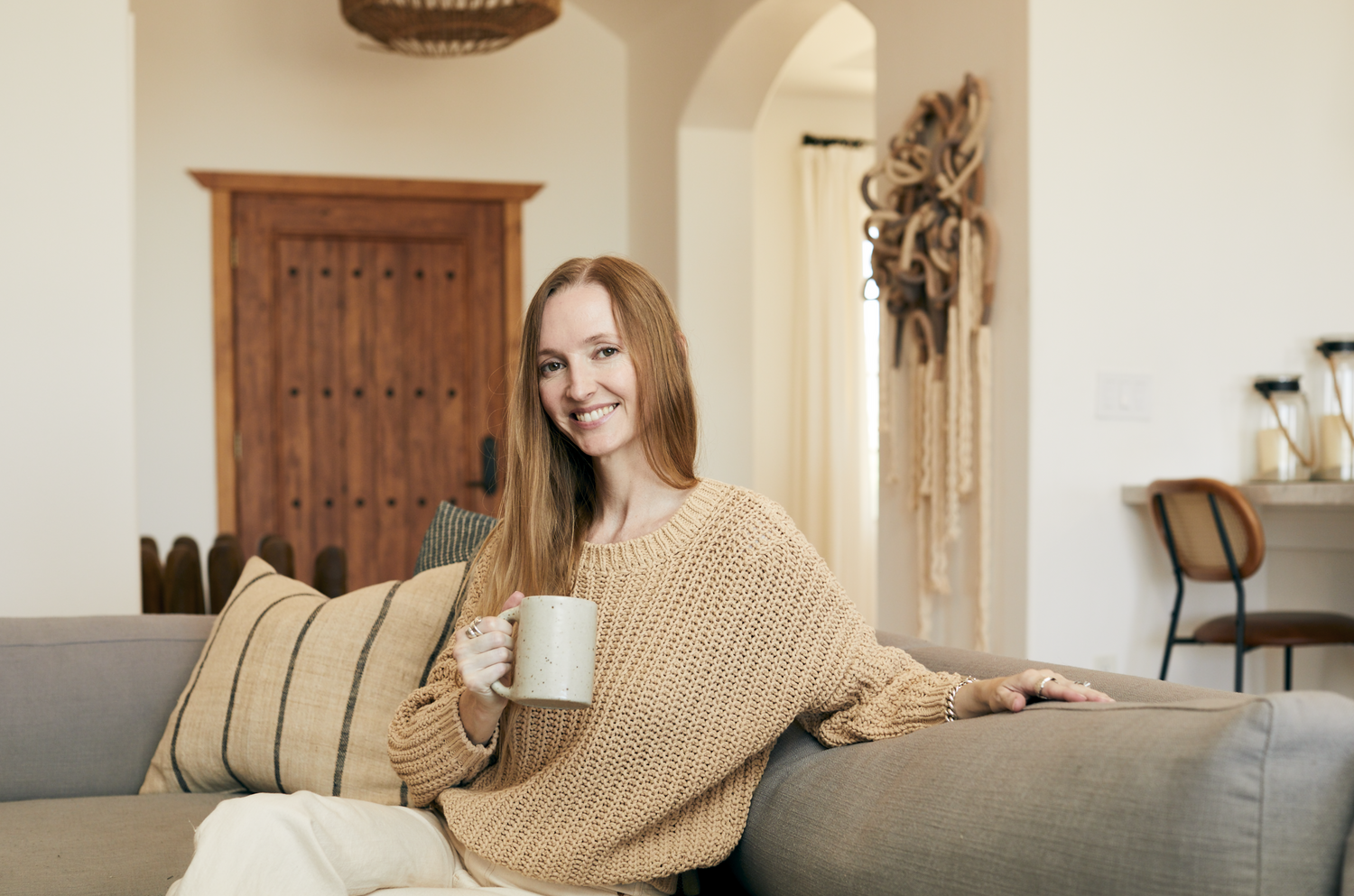 Woman relaxing on gray couch with coffee mug in cozy living room with arched doorway.