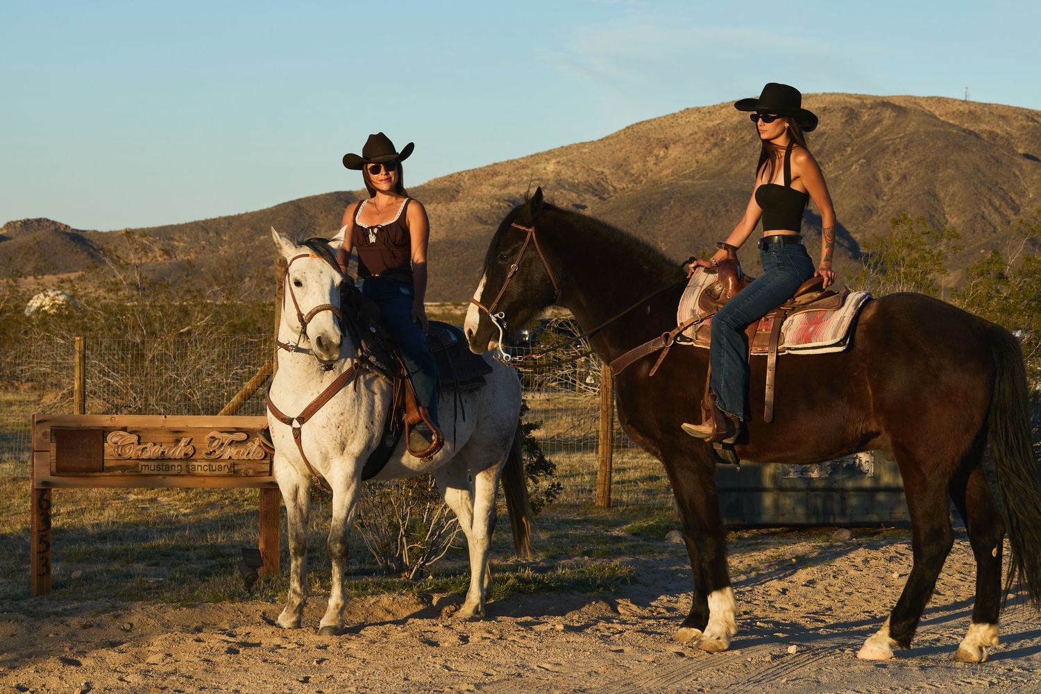 Two cowgirls in black hats ride a white and brown horse in a desert ranch setting at golden hour.