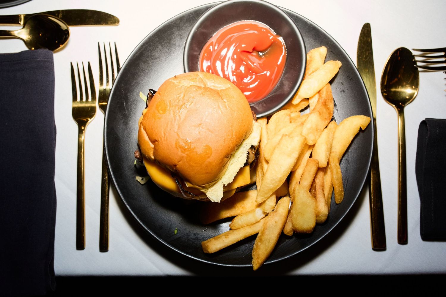 A cheeseburger and french fries served on a black plate with red dipping sauce and silverware on white tablecloth.