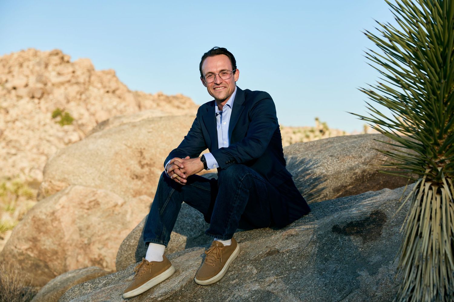 A person in business attire sits on a large boulder in a desert landscape with yucca plants and rocky terrain.
