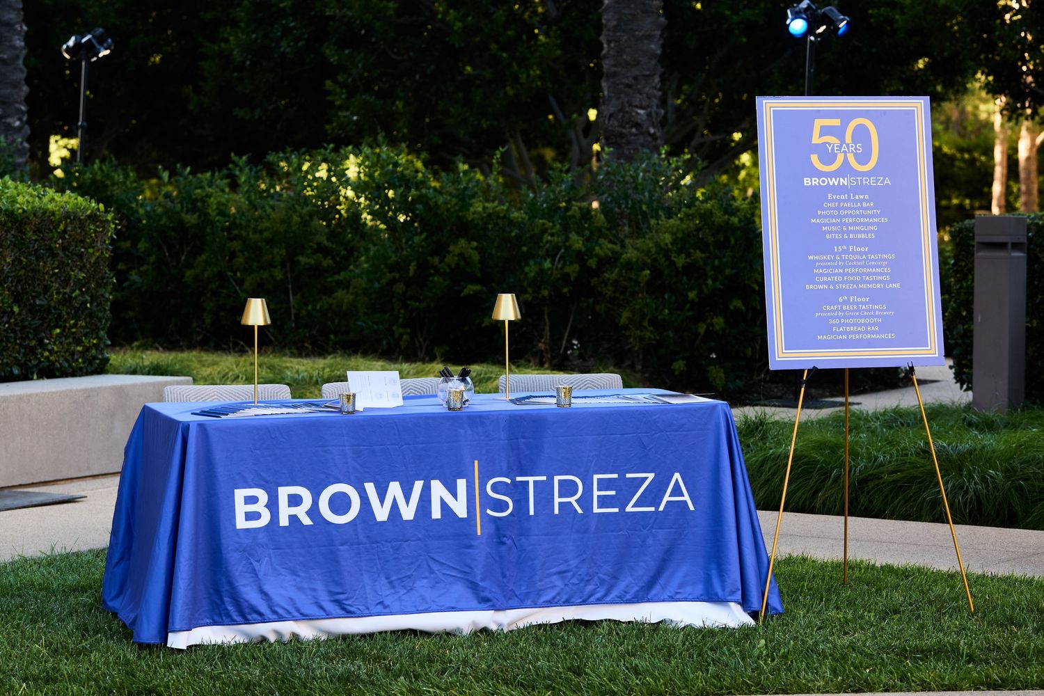 A series of outdoor event tables with blue tablecloths and signage set up in a garden area at dusk.