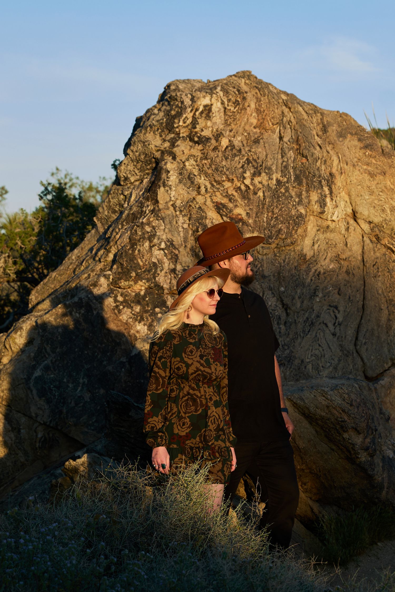 A couple stands before large rocks at golden hour, woman in patterned dress with sunglasses, man in brown cowboy hat.