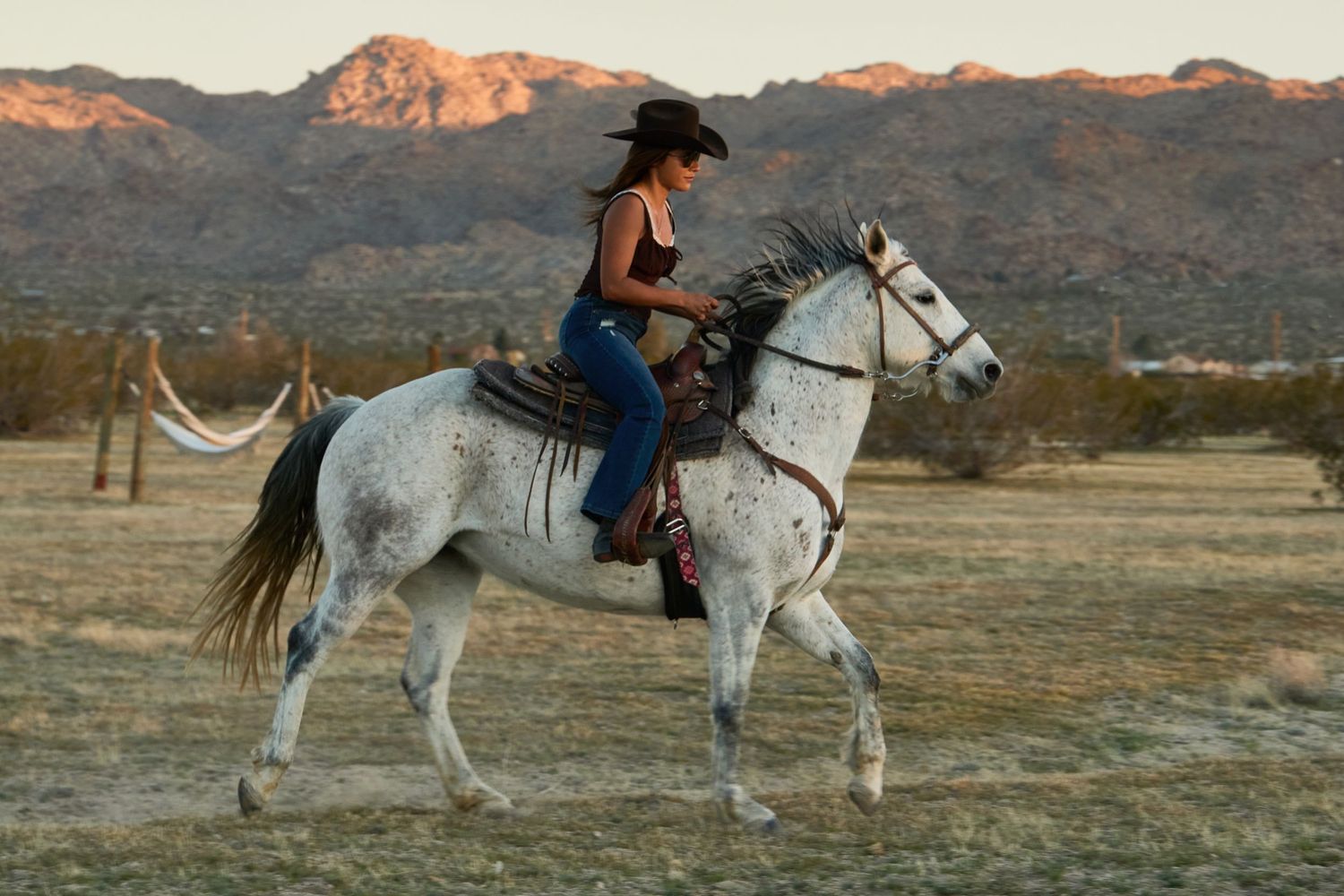 Woman in cowboy hat riding a white Appaloosa horse across desert landscape with mountain backdrop at golden hour.