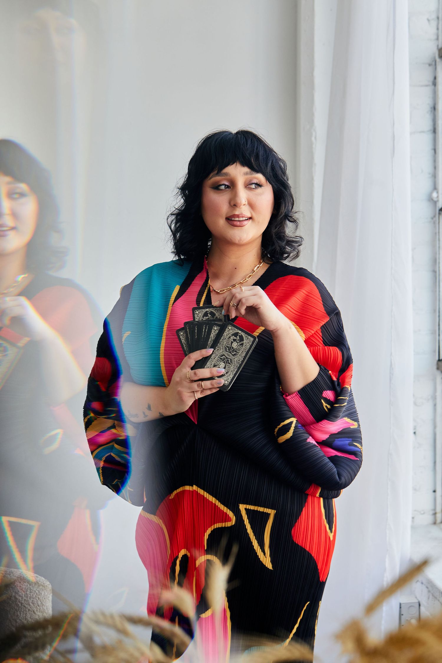 Woman in colorful geometric print dress holding tarot cards, posing in bright studio with reflection visible.