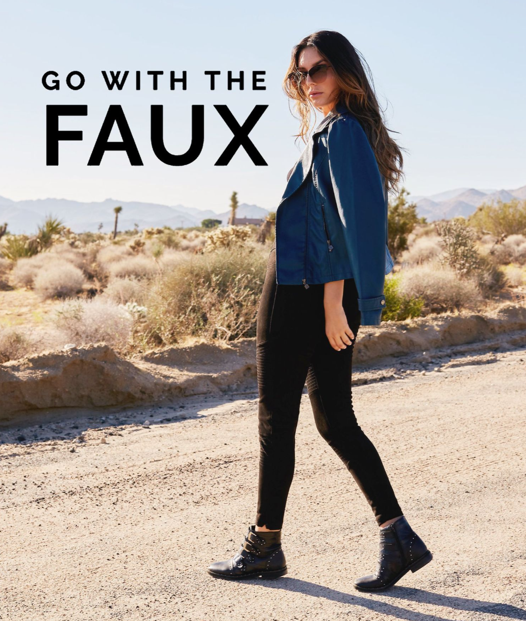 Person in faux leather jacket and black pants walking on desert path with mountains in background.