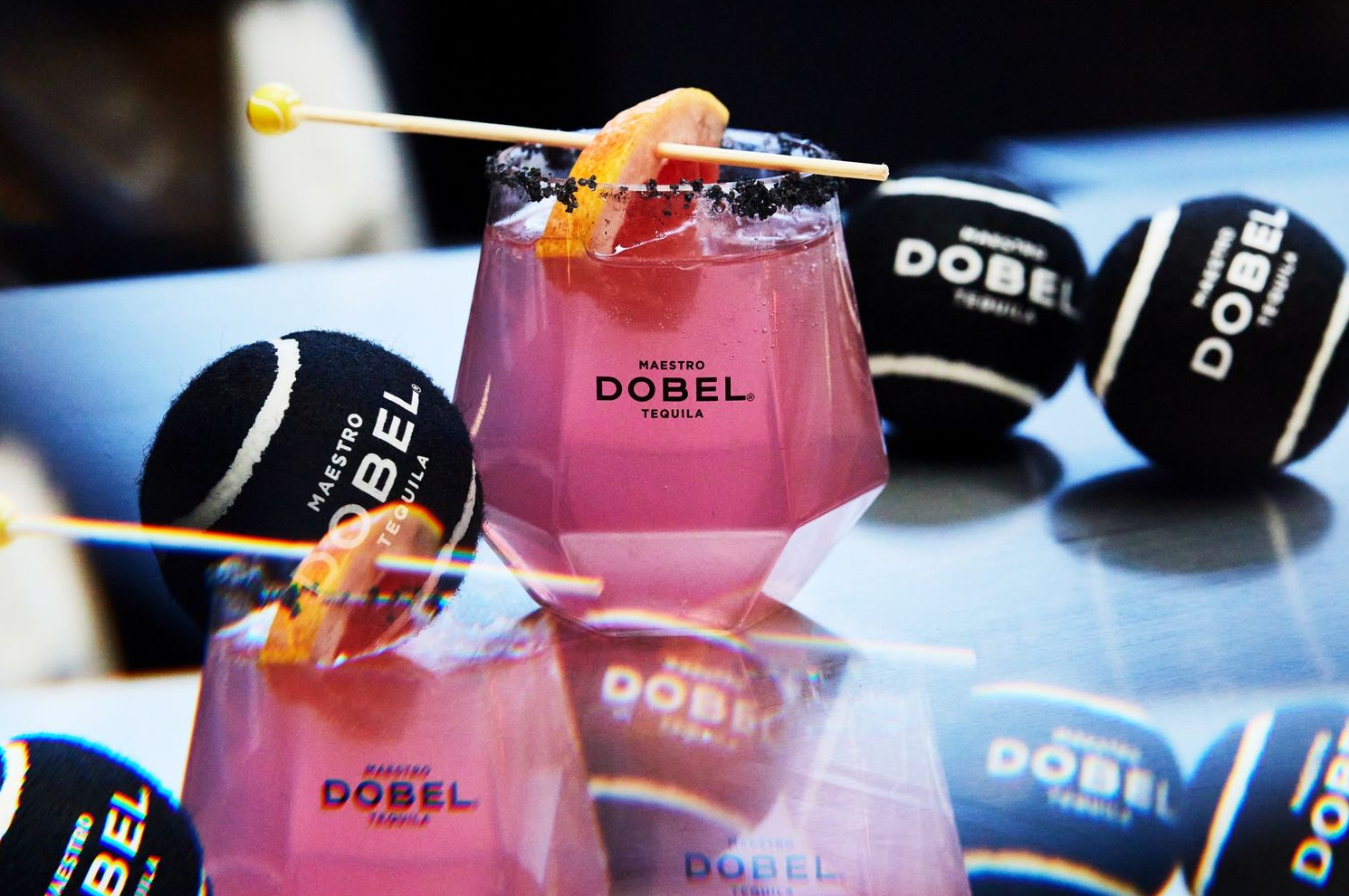 Pink cocktails with citrus garnish in branded Dobel glasses surrounded by black promotional items.