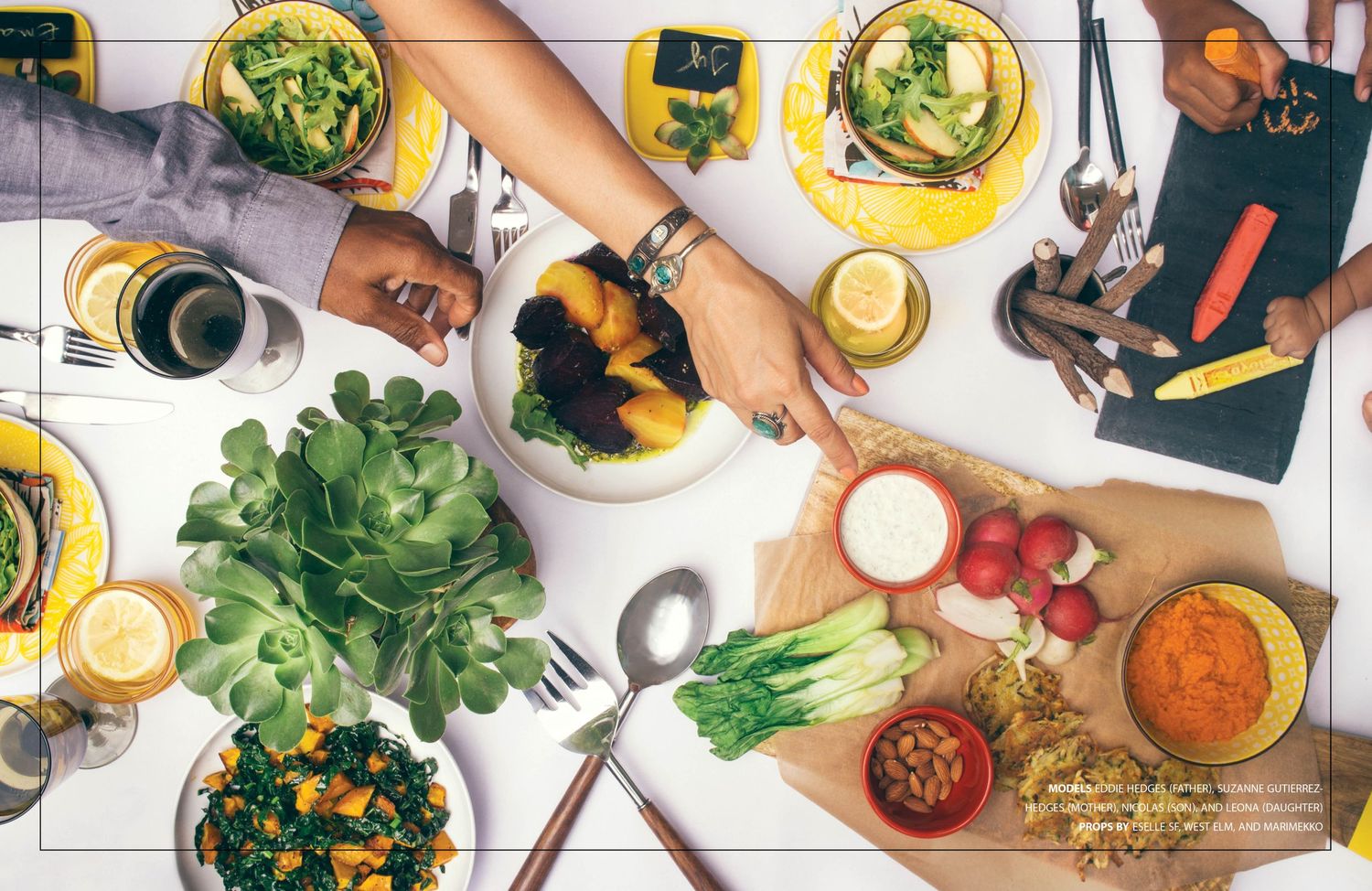 Overhead view of hands sharing a healthy meal with fresh produce, drinks and colorful dishes on a white table.