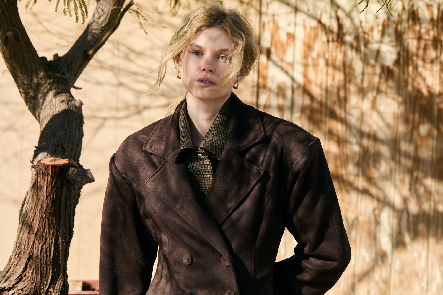 A person in a dark jacket poses against a beige wall with tree shadow patterns creating an artistic composition.