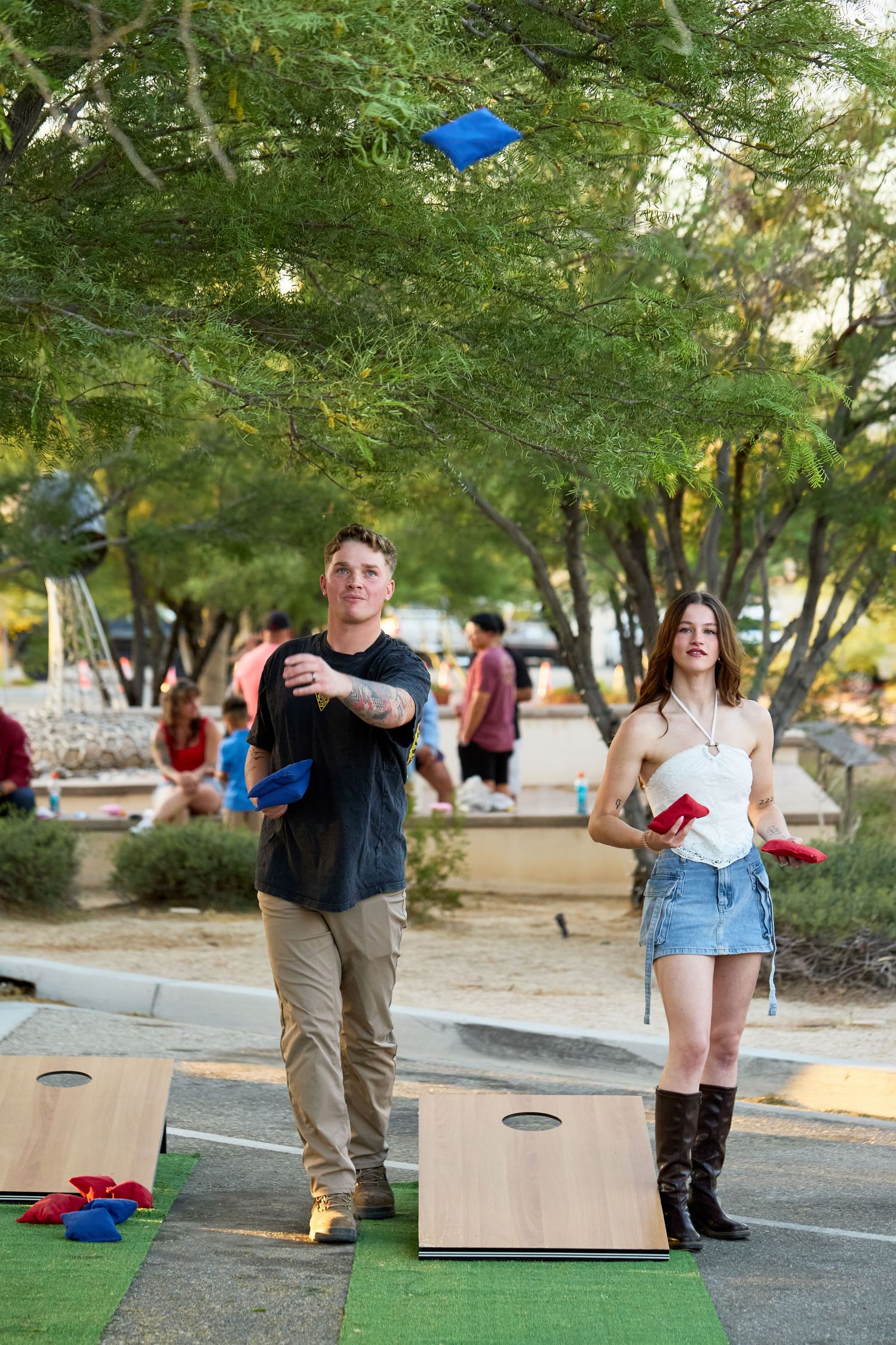 Two people playing cornhole outdoors in a park, man tossing blue bag while woman watches, trees in background.