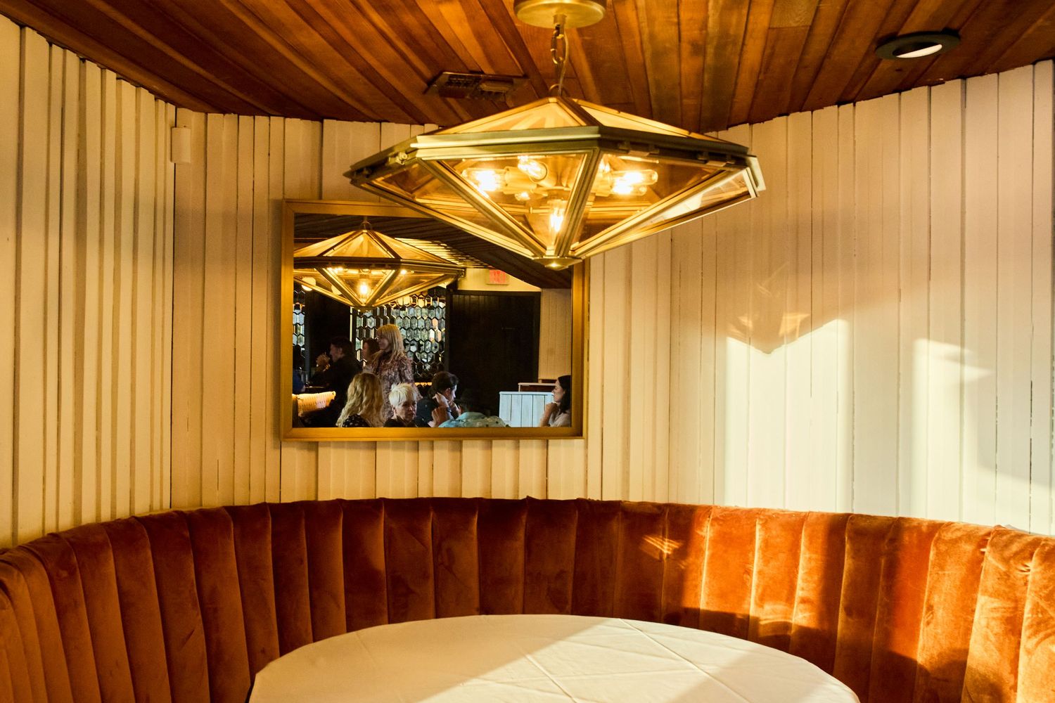 A modern restaurant booth with orange velvet seating, wooden ceiling panels, and geometric gold light fixture.