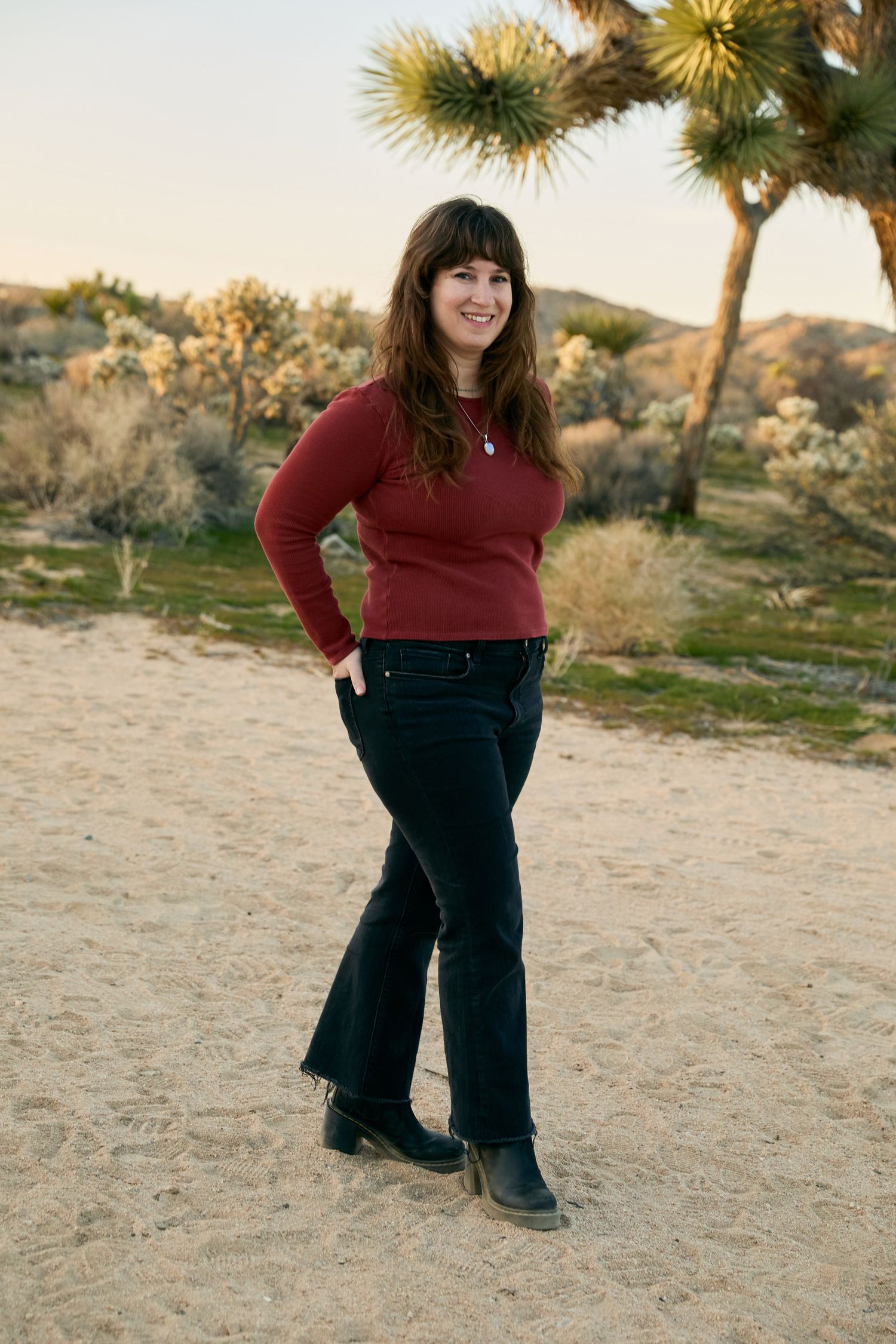 A person in a burgundy sweater and dark jeans stands on sandy ground with a Joshua tree in the desert background at sunset.