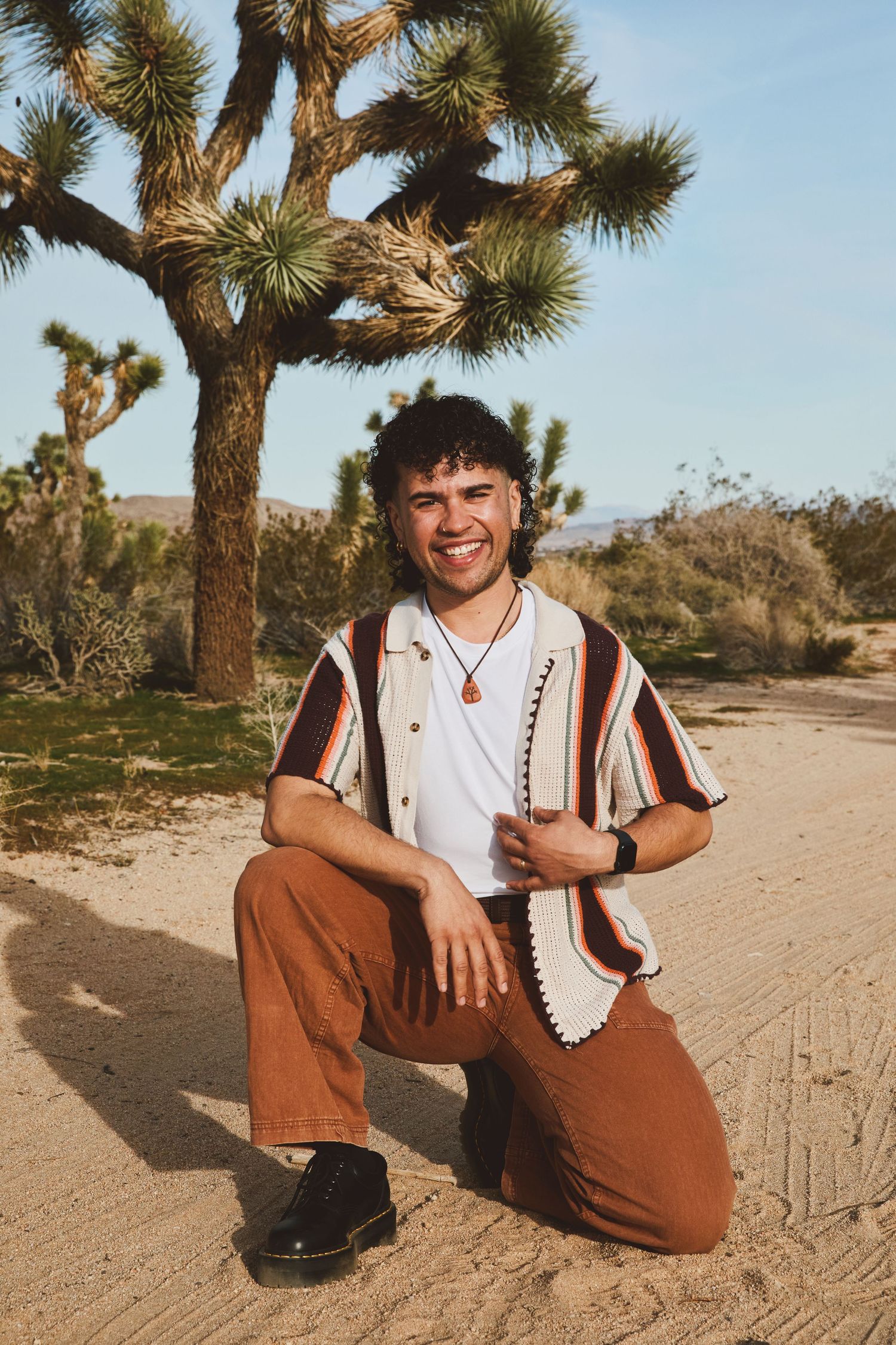 A person poses in casual attire near a Joshua Tree in a desert landscape during golden hour.