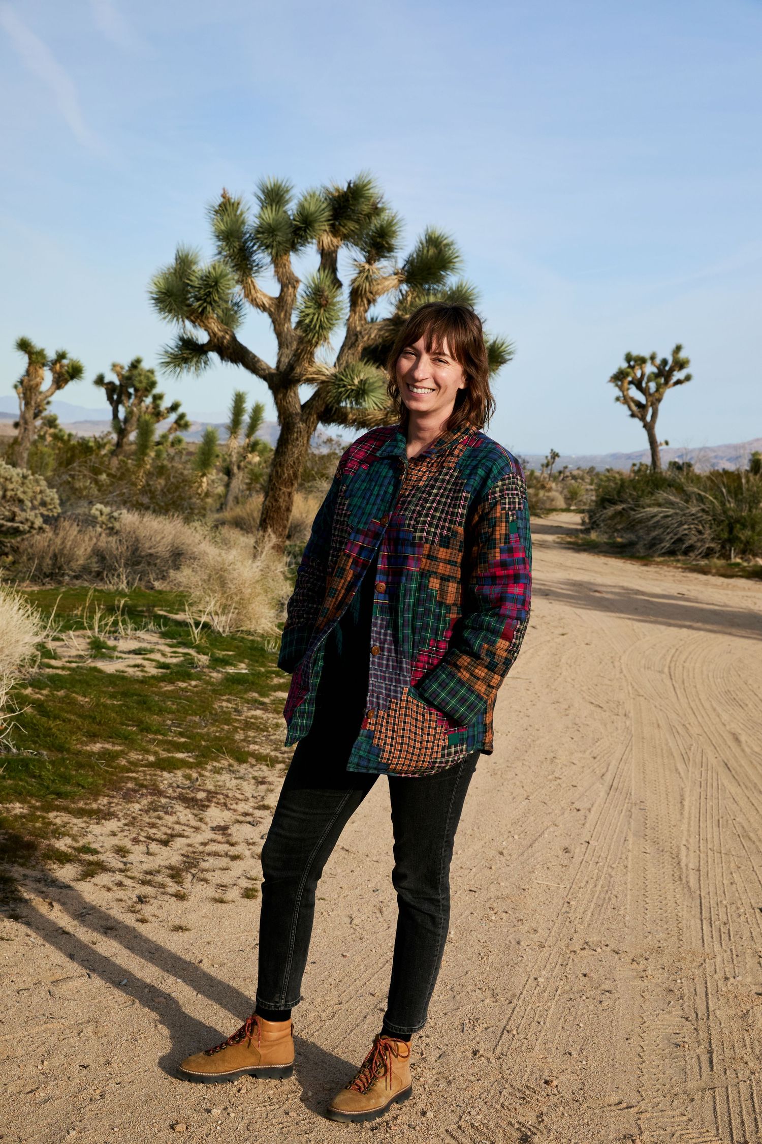 Woman smiling in colorful plaid jacket, black jeans, and boots on a desert dirt road with Joshua trees.