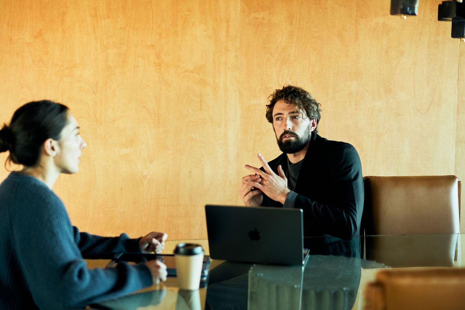Two people having an animated business conversation at a table with a laptop and coffee in an office setting.