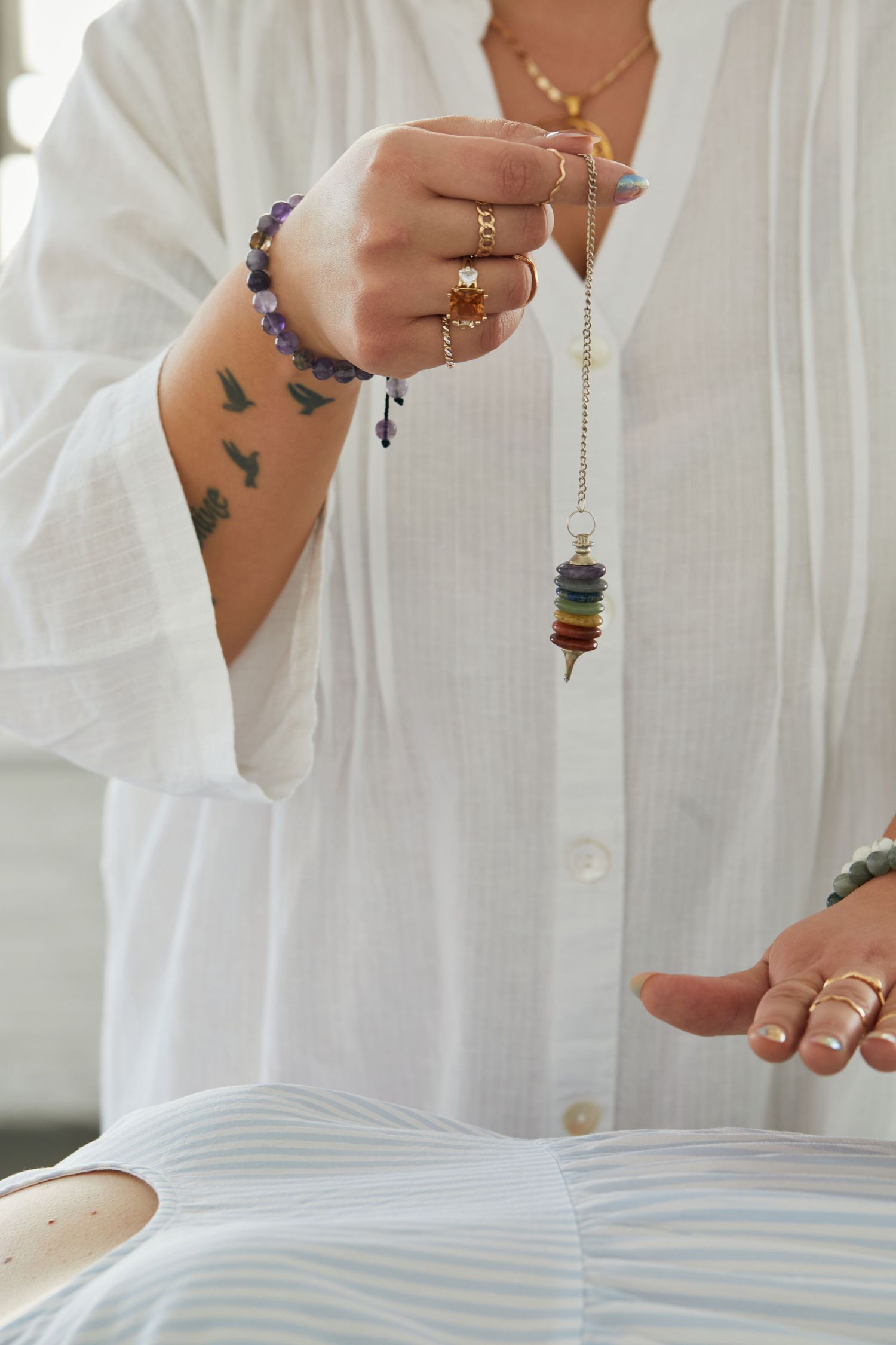 Woman holding a chakra crystal pendulum pendulum with tattoos, rings, and amethyst bracelet in white linen shirt.