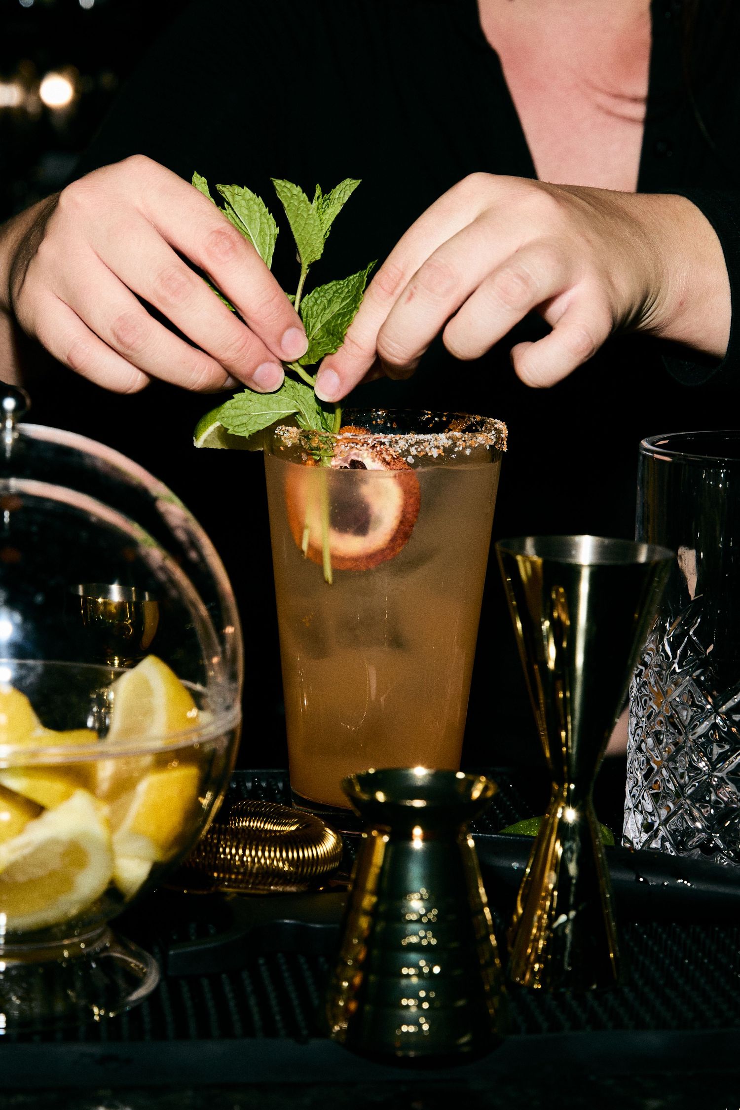A bartender garnishes a cocktail with fresh mint leaves next to sliced lemons and bar tools on a dark counter.
